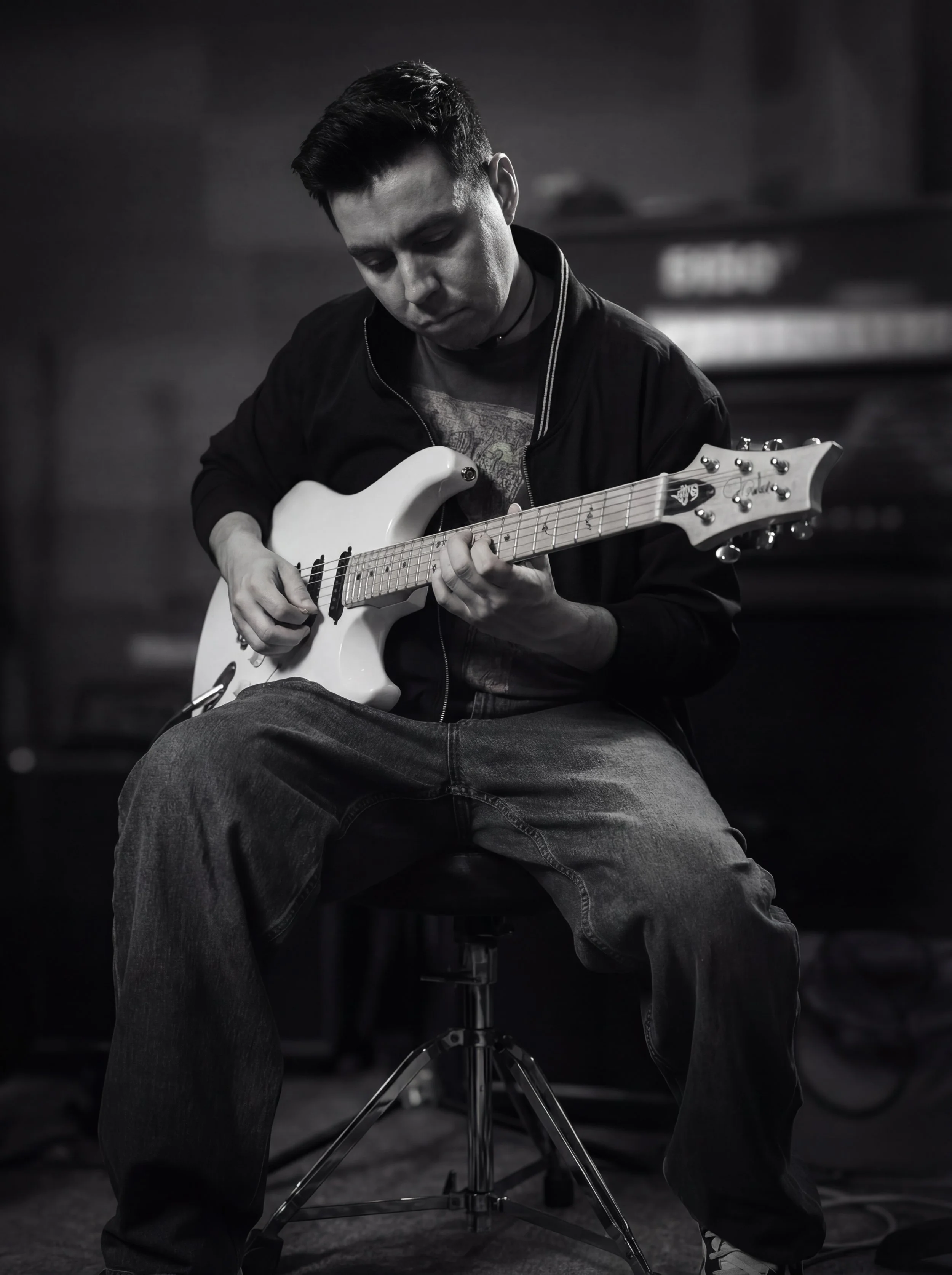 A young man playing an electric guitar while sitting on a stool, in a black and white photo with a blurred background.