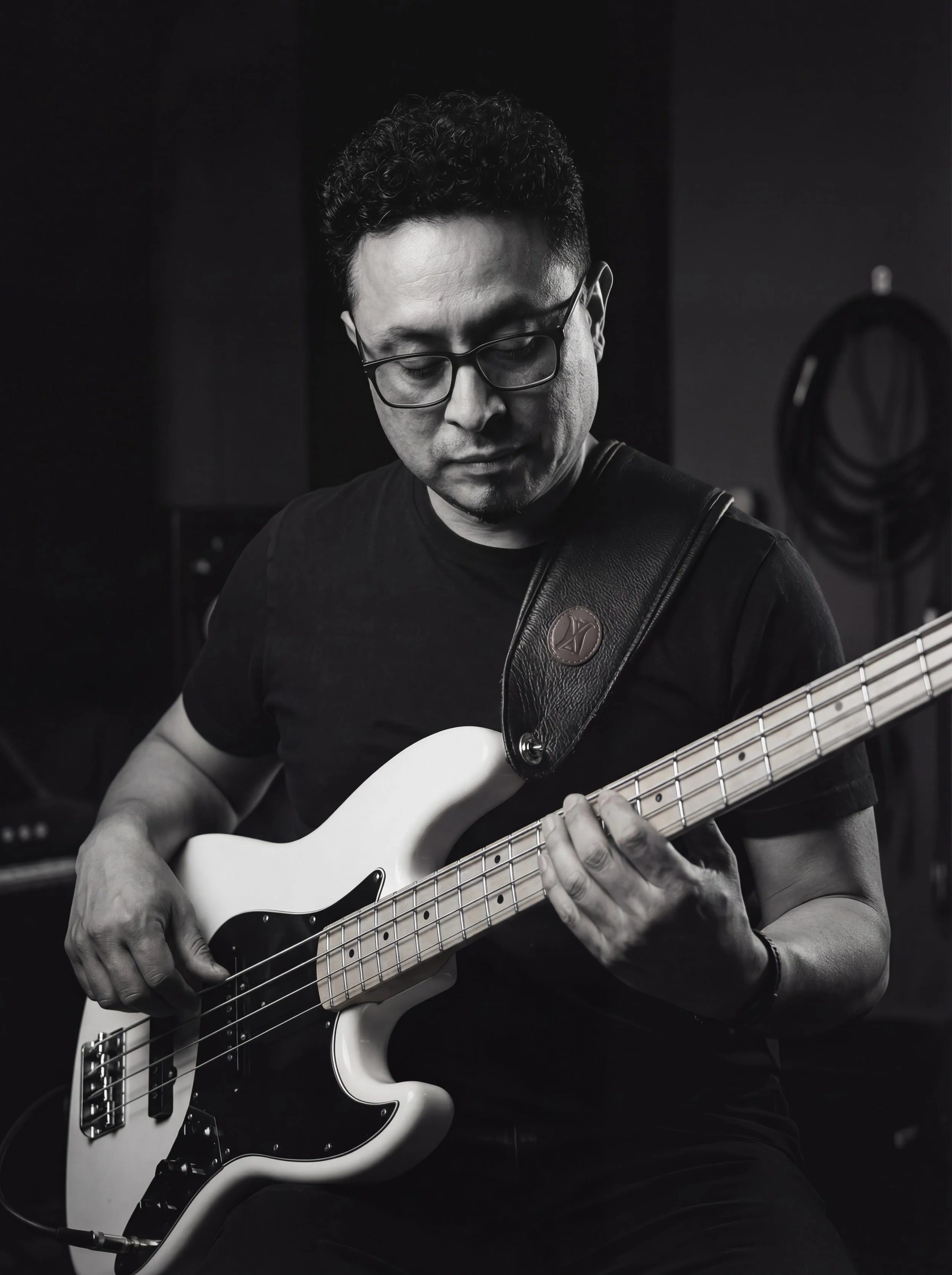 A man with short curly hair, glasses, and a black t-shirt playing a white electric bass guitar in a recording studio.