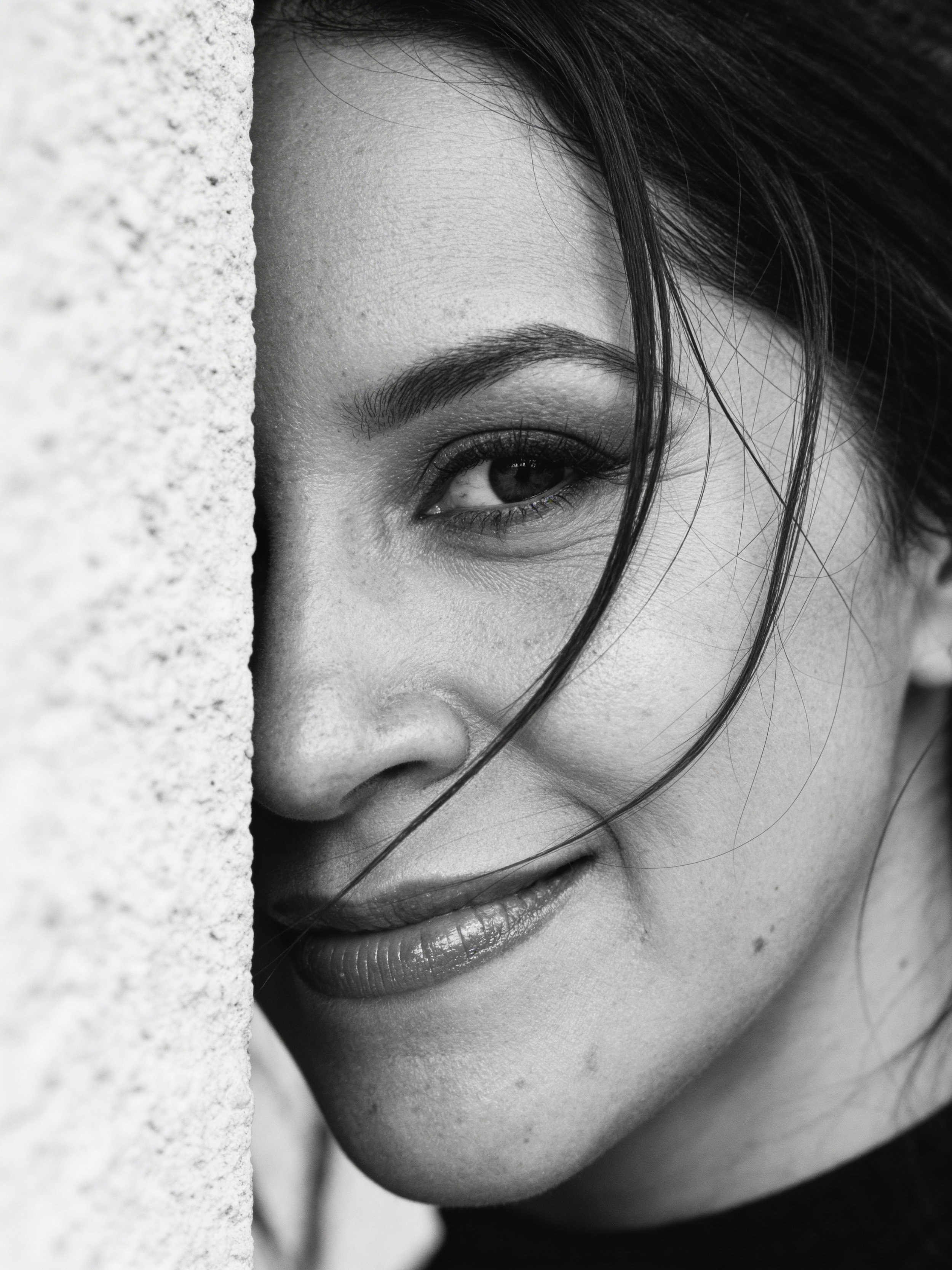Close-up black and white photo of a woman's face peeking from behind a wall, with only one eye and part of her lips visible, highlighting her eye and facial features.