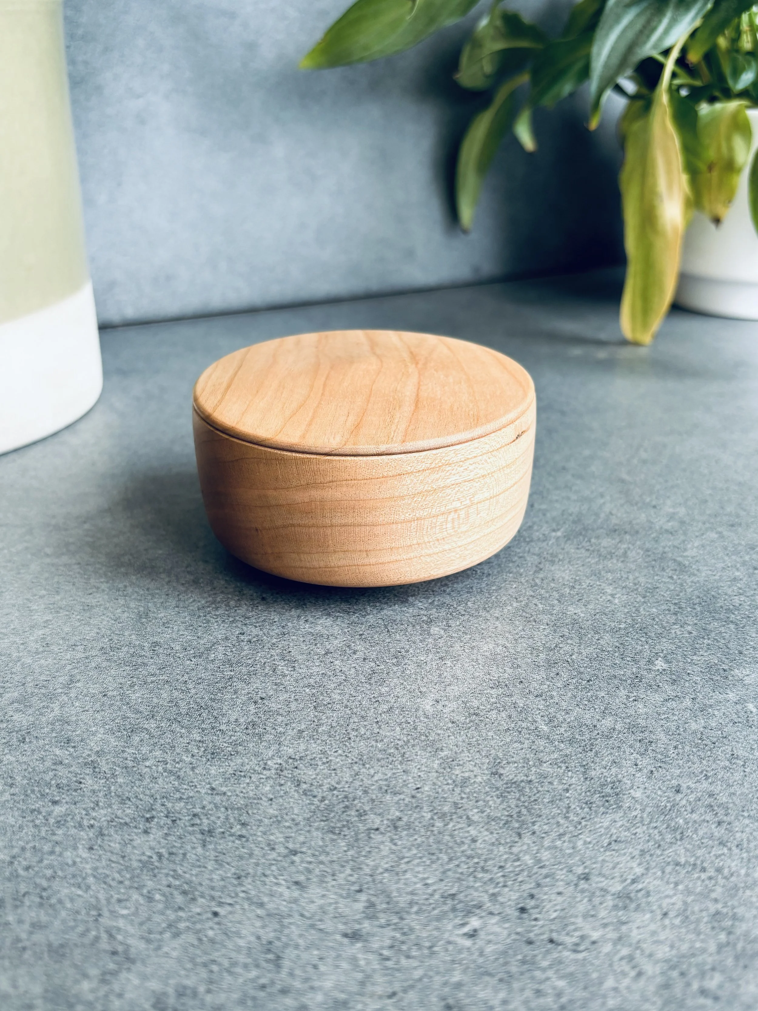 A round wooden container with a lid on a gray surface, with green plants in the background.