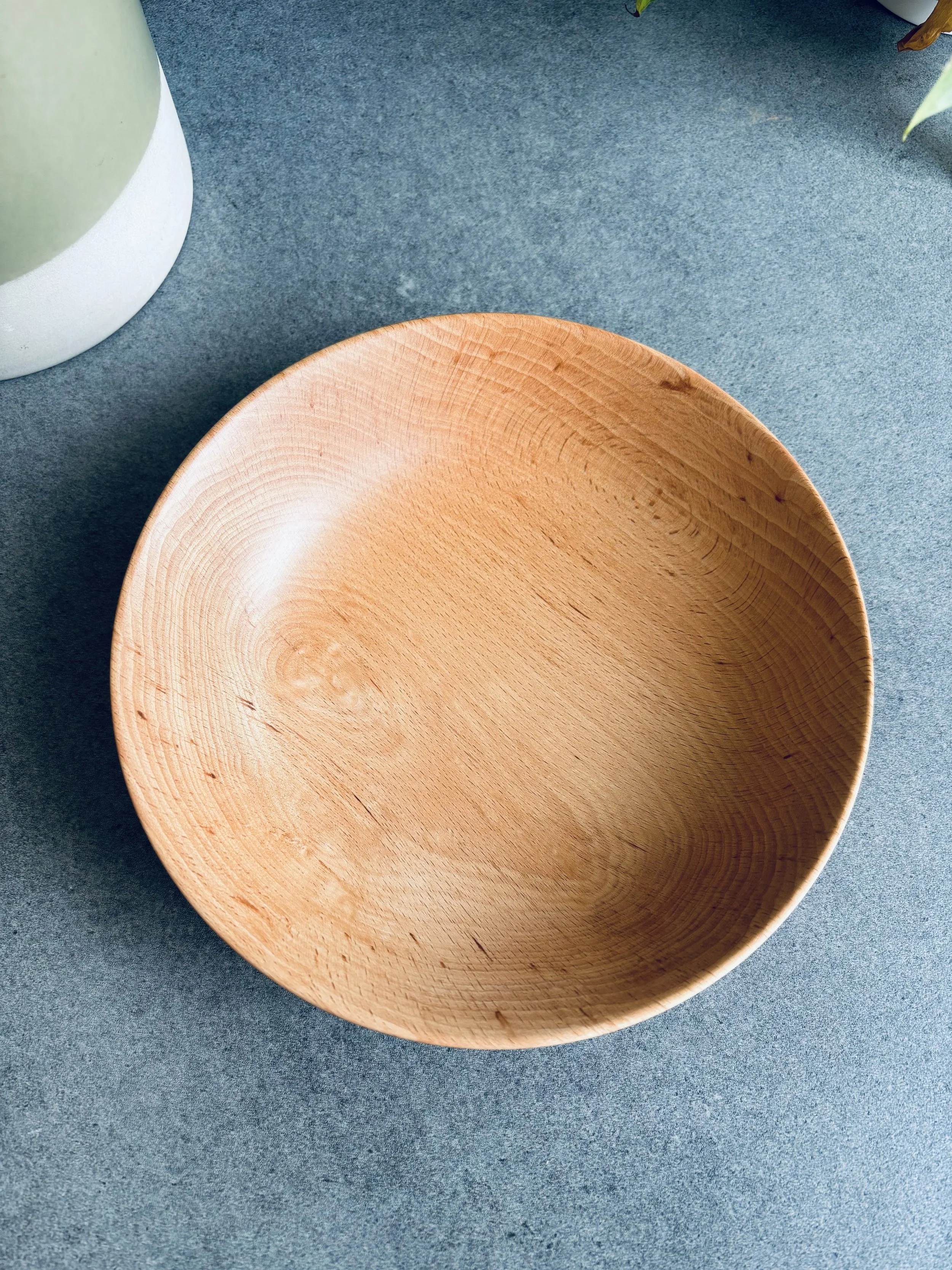 Empty wooden bowl on a blue surface with a partial view of a green and white pot to the upper left and some green leaves to the upper right.
