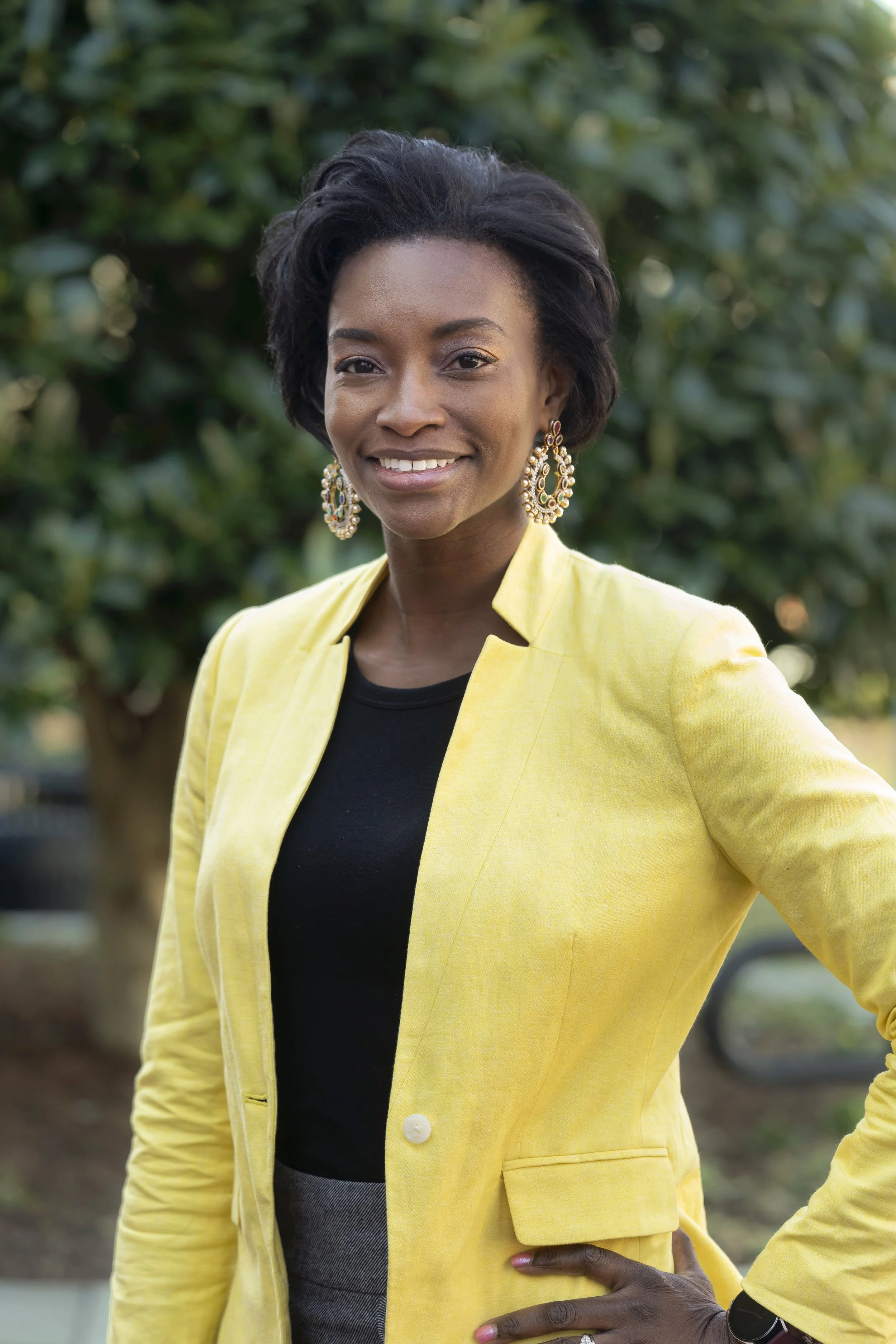 A smiling Black woman standing outdoors in a garden or park setting, wearing a yellow blazer, a black top, and large, ornate earrings.