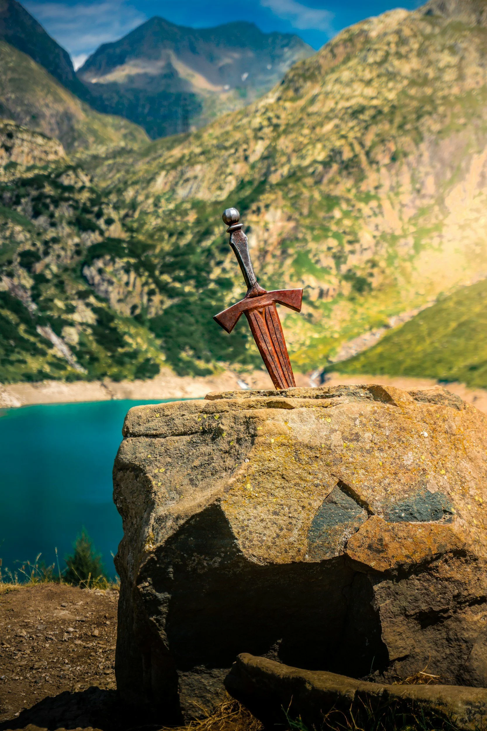 A small wooden sword stuck into a large rock overlooking a mountain lake and green foothills.