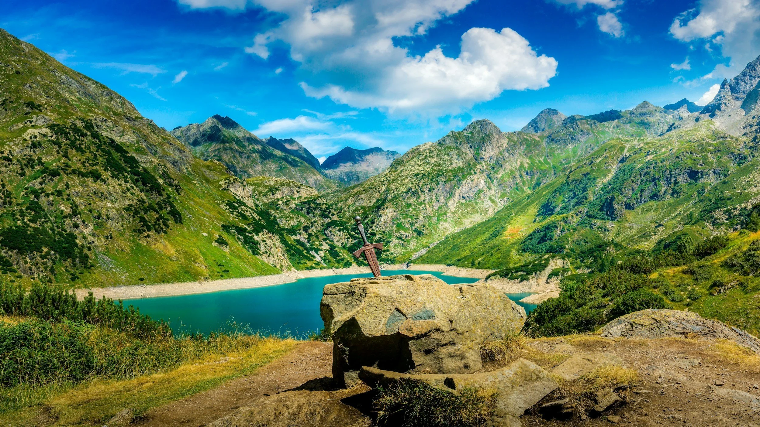 A scenic mountain landscape with green slopes, a blue lake, and a partly cloudy sky. In the foreground, a large rock has a sword embedded in it.