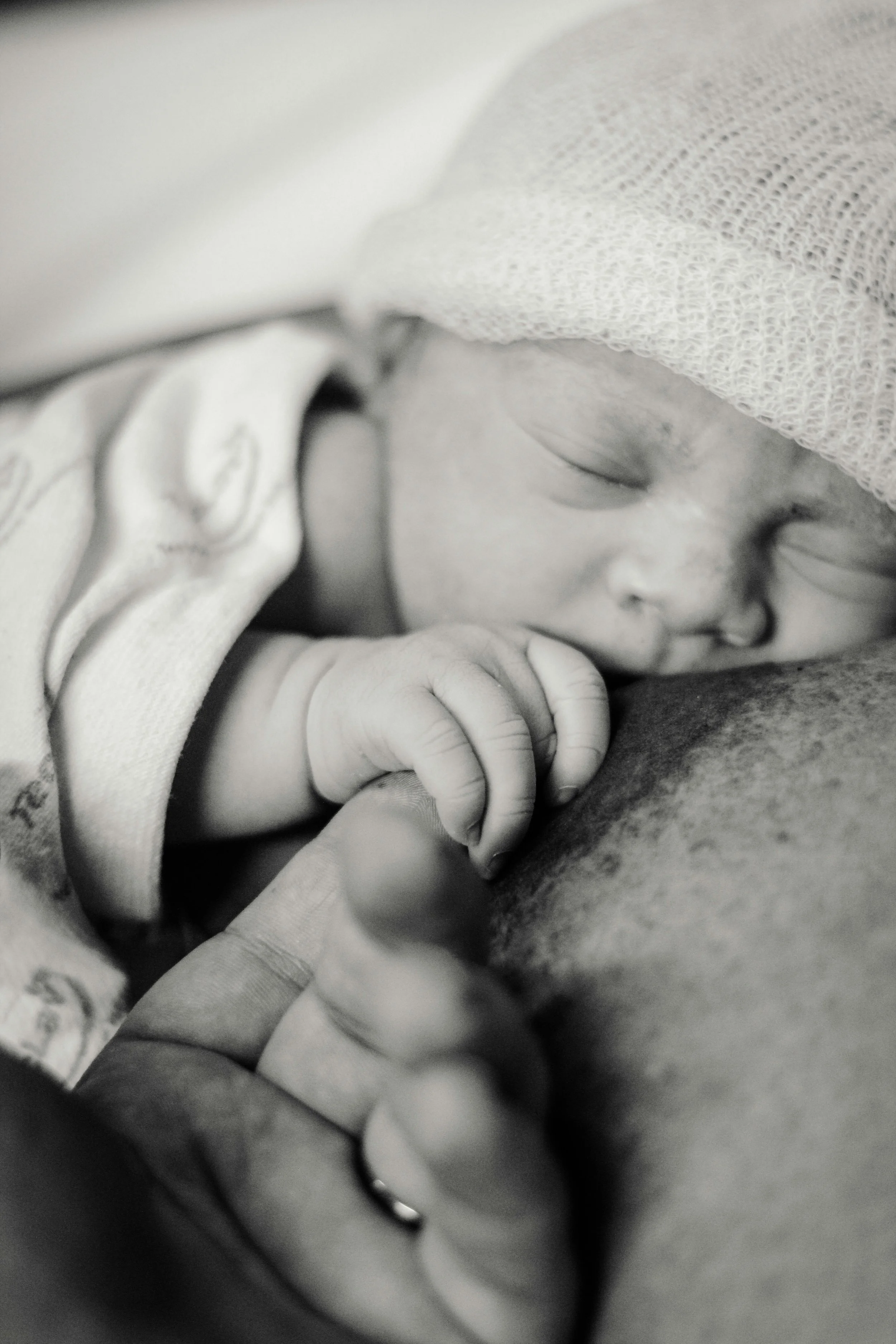 Close-up black and white photo of a newborn baby sleeping, with a surgical cap on its head and hands gently touching its face.