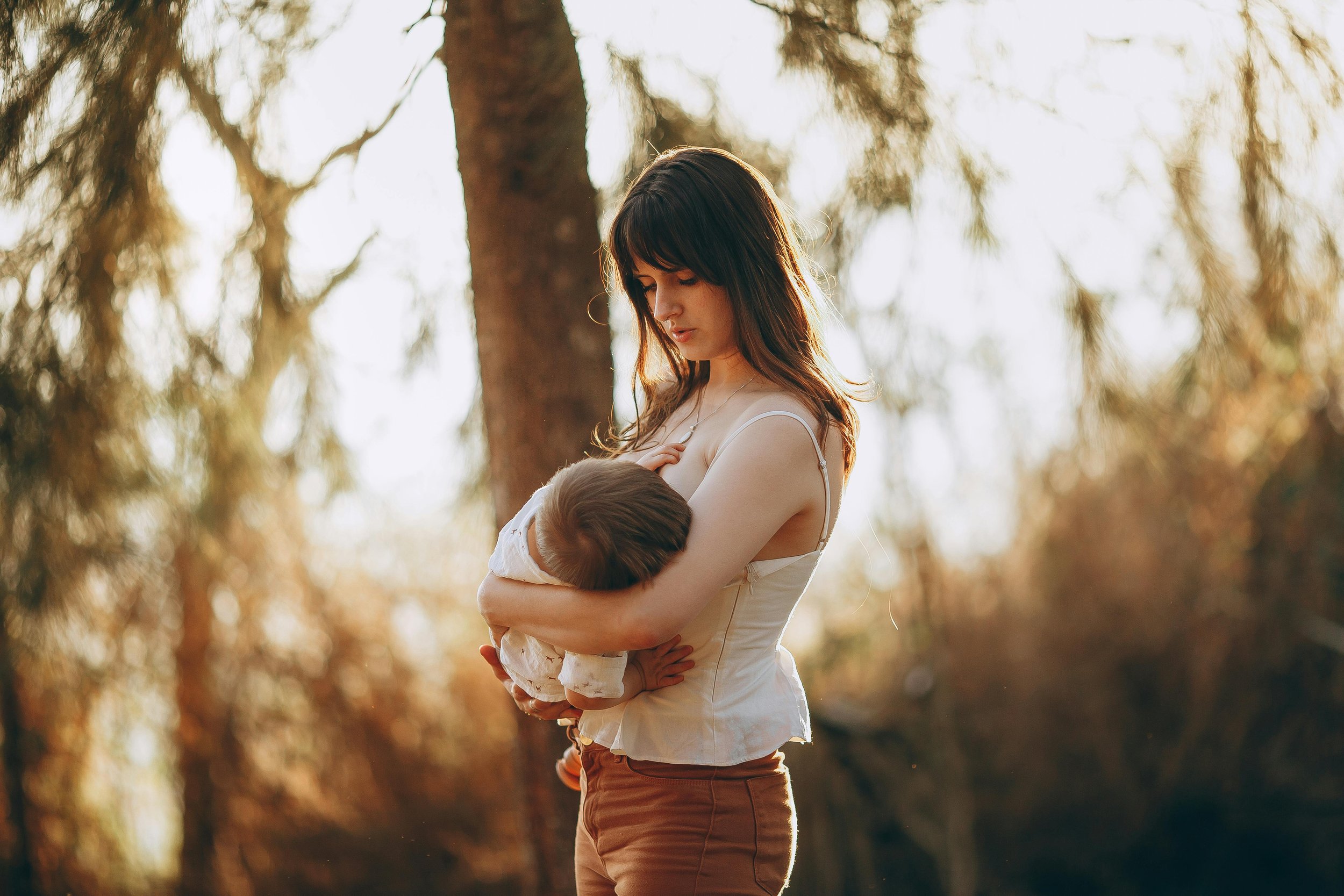 A woman holding a child in her arms outdoors in a forested area with sunlight filtering through the trees.