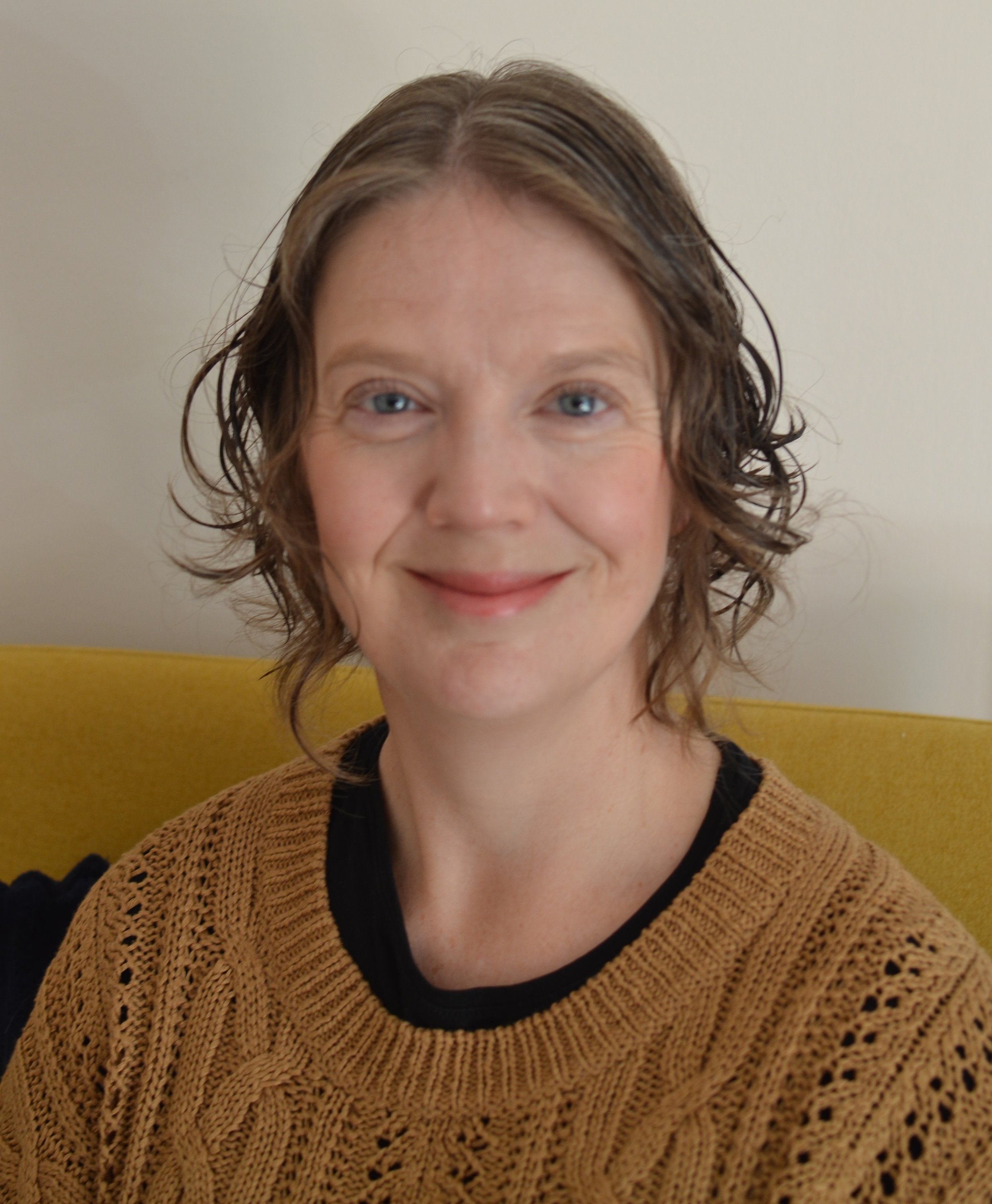 A woman with wavy, shoulder-length hair smiling, wearing a black shirt and a brown knit cardigan, sitting against a plain light-colored wall with a yellow cushion behind her.