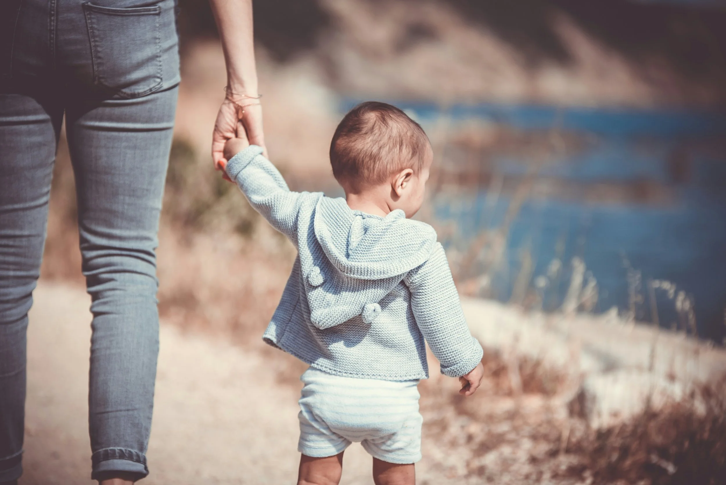 A young child holding an adult's hand while walking outdoors near a body of water, with the adult wearing gray jeans and the child in a gray hooded sweater and striped shorts.