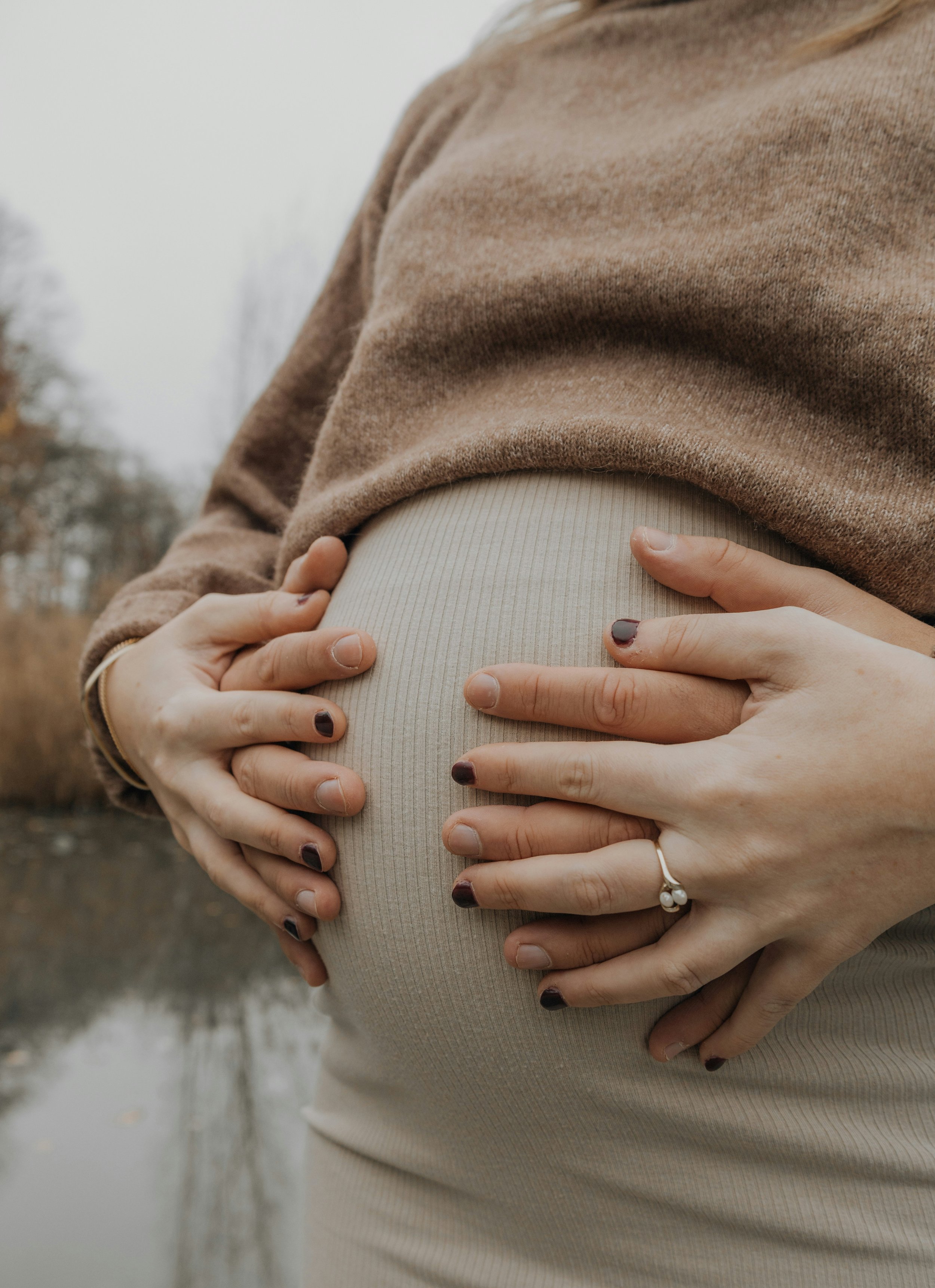 A pregnant woman wearing a tan sweater and beige ribbed leggings, with hands resting on her belly outdoors.