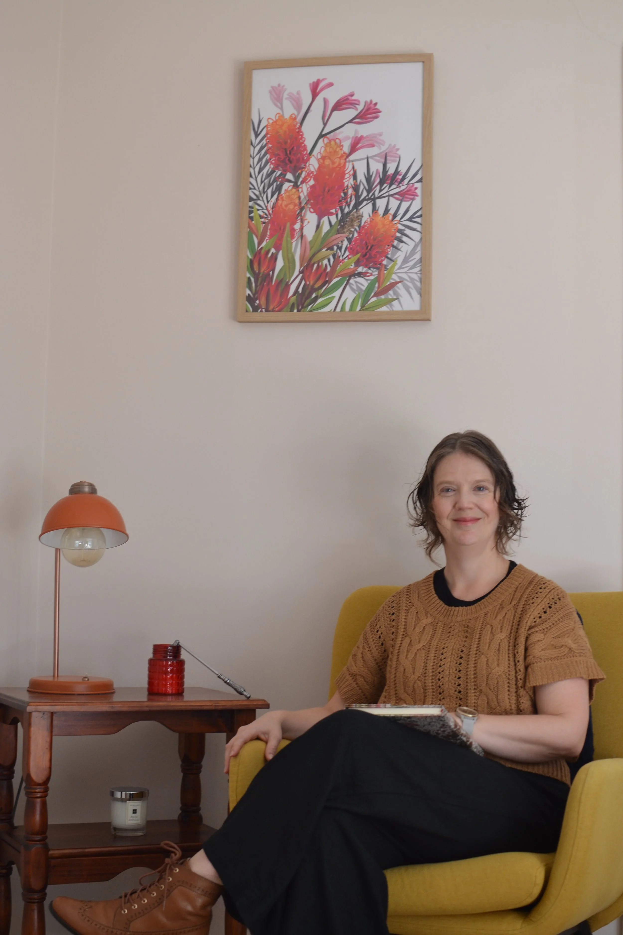 A woman with curly hair wearing a brown sweater sitting in a yellow armchair in a room with a picture of pink, orange, and purple flowers on the wall, a wooden side table with a orange lamp and a red jar, and a small object on the floor.