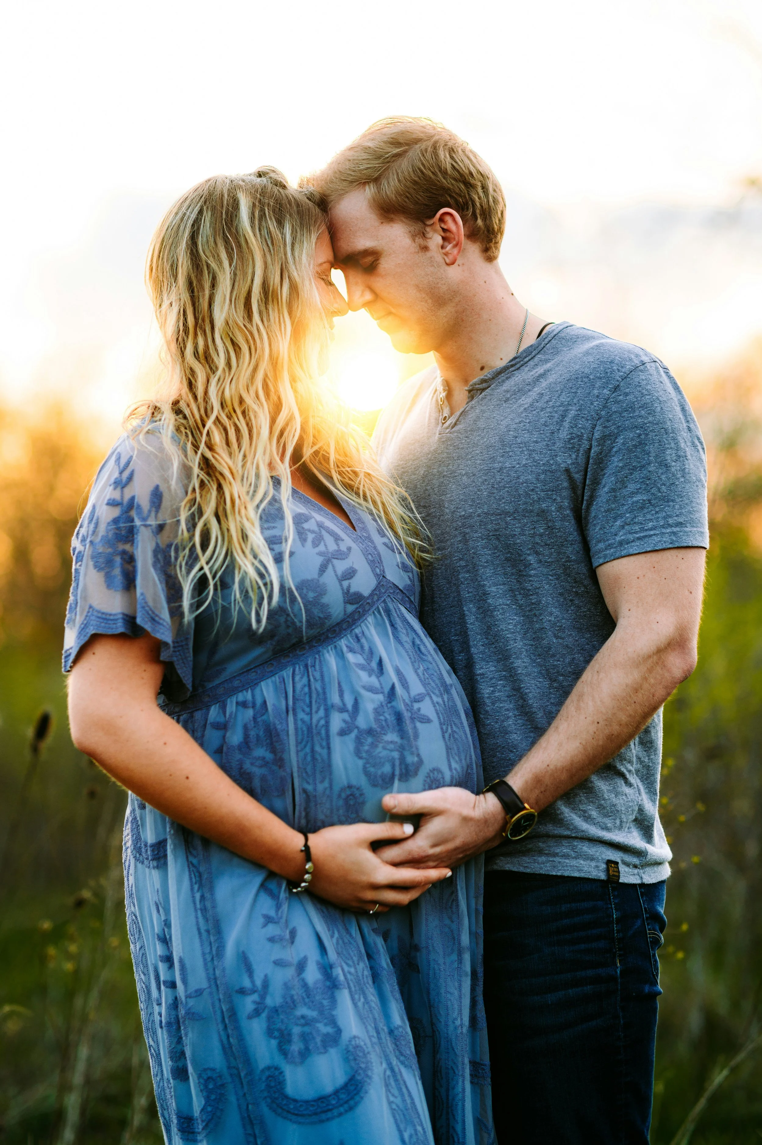 A pregnant woman and a man are standing outdoors, touching foreheads with eyes closed, in a warm embrace during sunset.