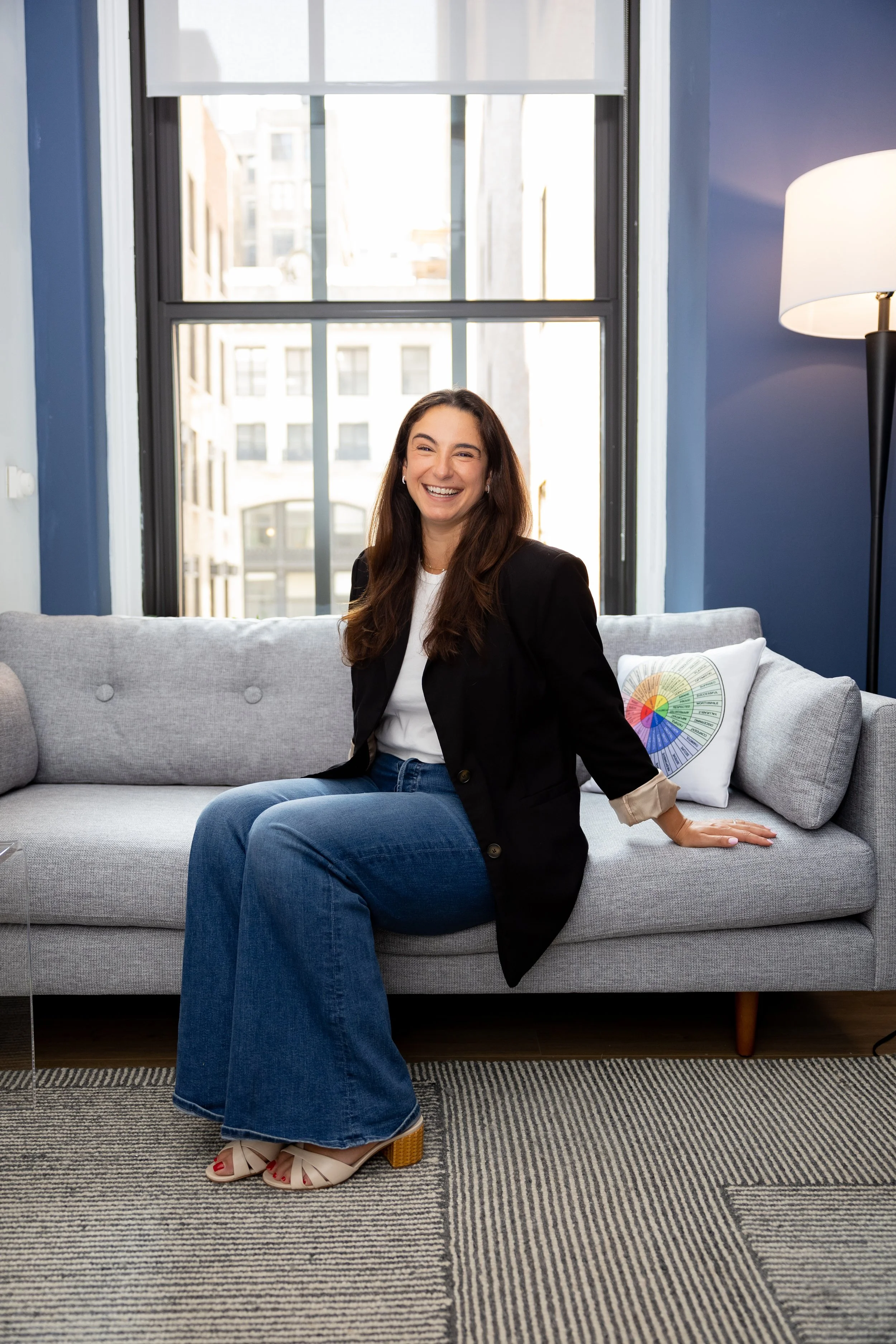 A Therapist with long brown hair, wearing a white shirt and black blazer, sitting on a light gray sofa in a modern room with large windows, a colorful wheel pillow, beige open-toe shoes with a chunky heel, and a dark blue wall with a tall floor lamp.