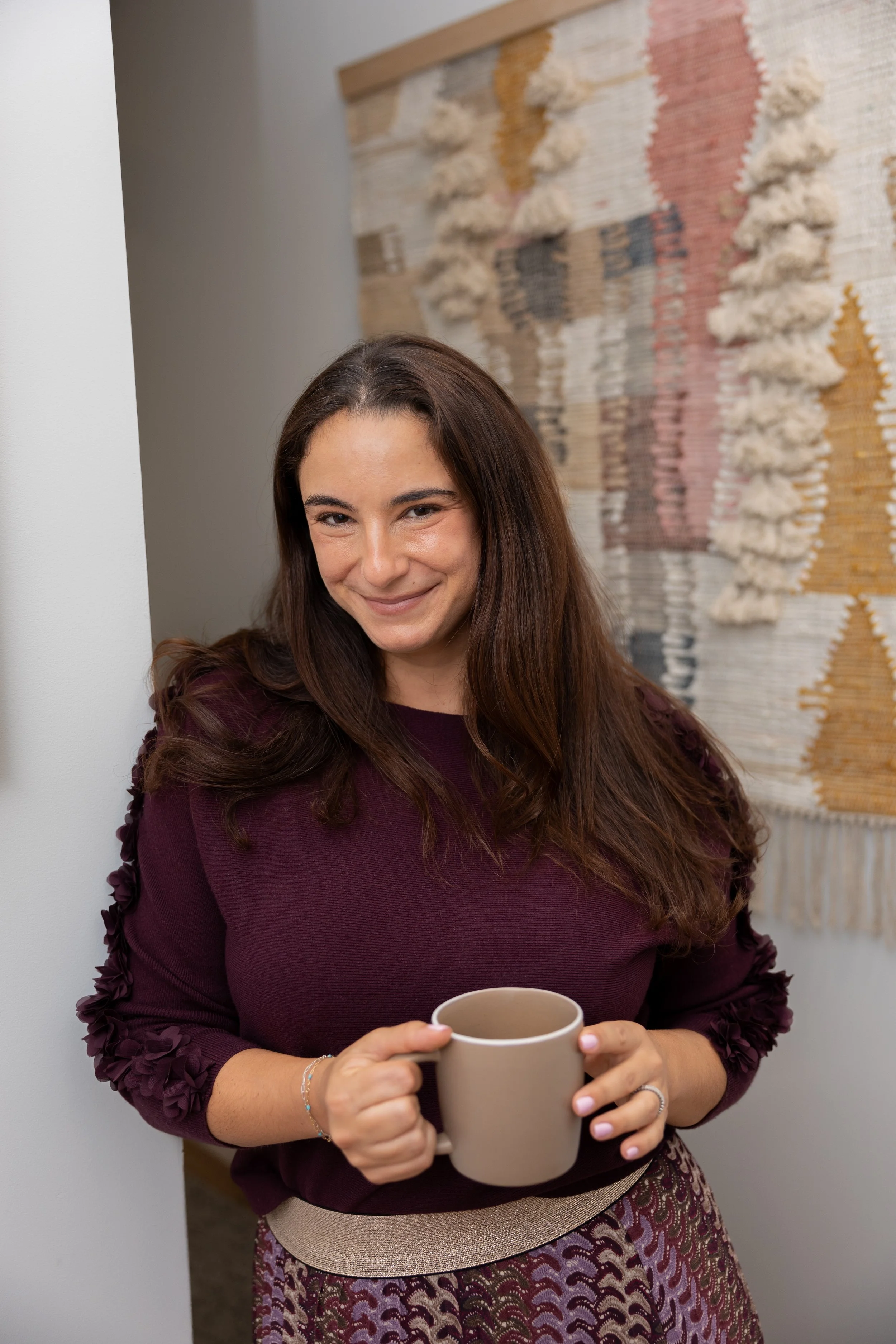 Therapist with long brown hair smiling while holding a beige coffee mug, standing in front of a textured woven wall hanging with geometric patterns.