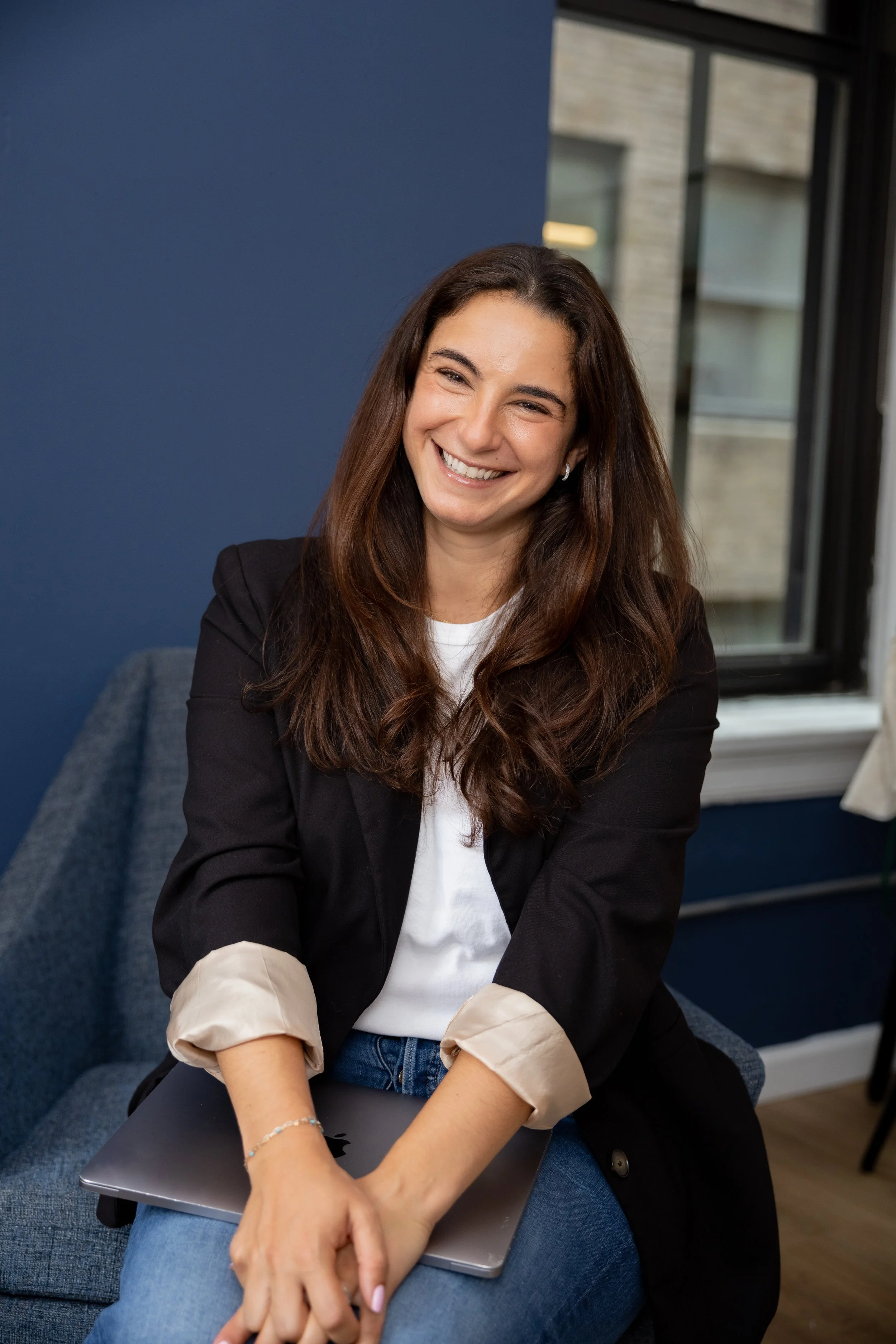A Therapist with long brown hair smiling, sitting on a blue chair, with a laptop on her lap, wearing a black blazer over a white shirt and light-colored rolled-up sleeves. There are large windows and a blue wall in the background.