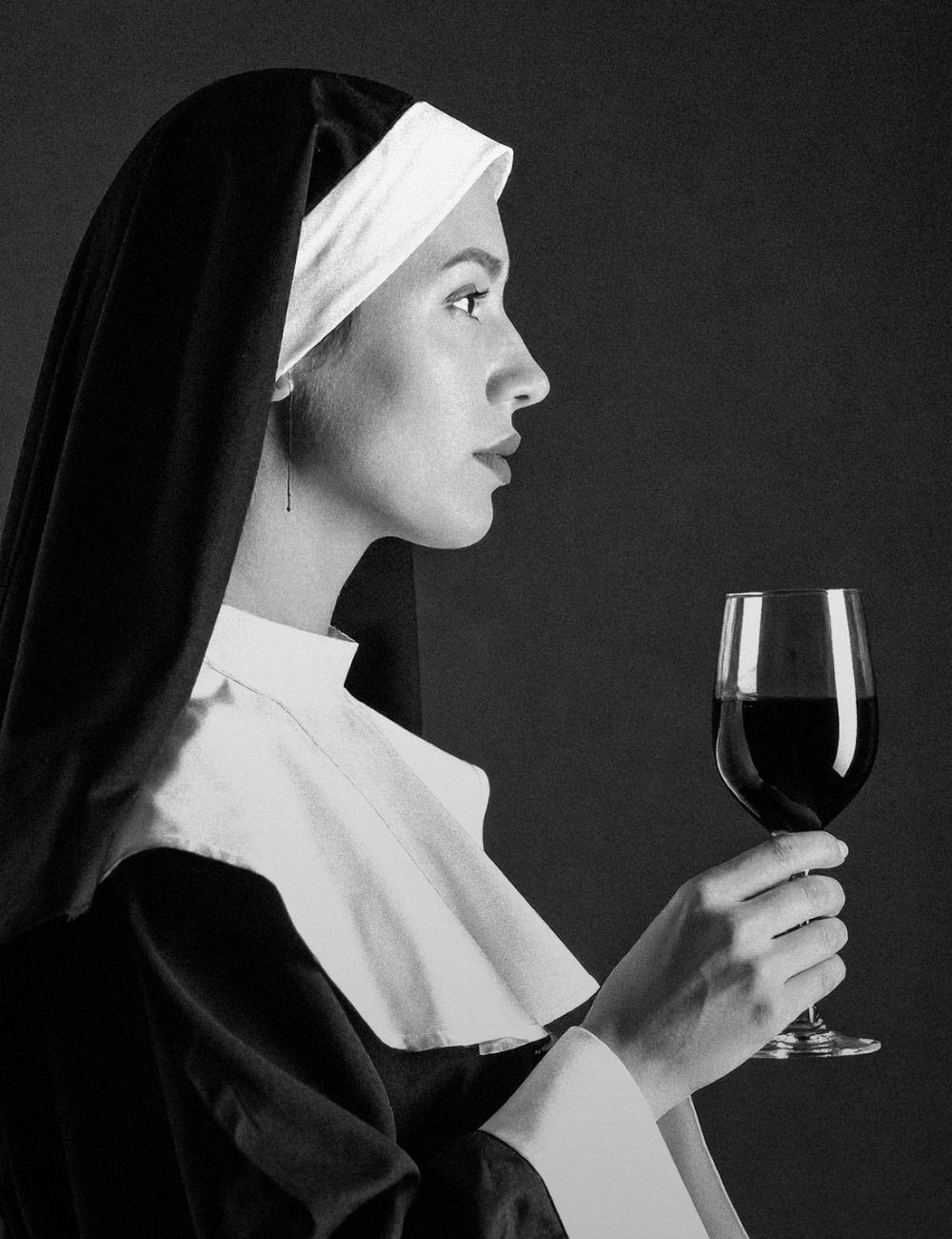 A black and white photo of a woman dressed as a nun holding a glass of red wine in her right hand, with her profile visible.