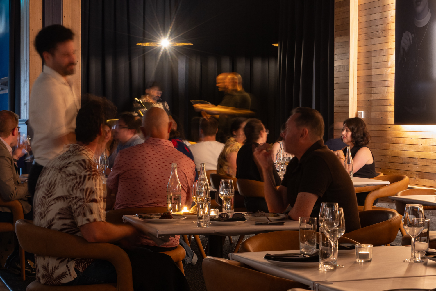 People dining at VICE restaurant with a live performance on stage, dim lighting, and wooden interior walls.