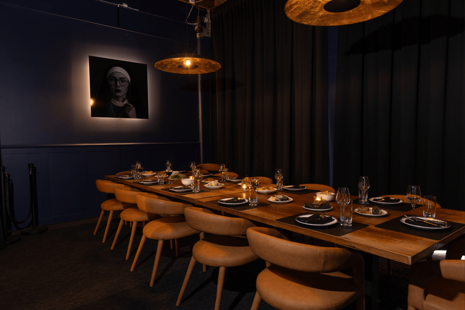A dimly lit VICE private dining room with a long wooden table set for a meal, with plates, glasses, and small candles, surrounded by beige chairs, with dark curtains on one side and a framed black-and-white portrait on the wall.