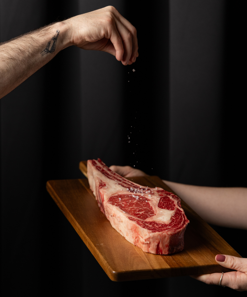 Person seasoning a raw steak with salt, on a wooden cutting board at VICE restaurant.