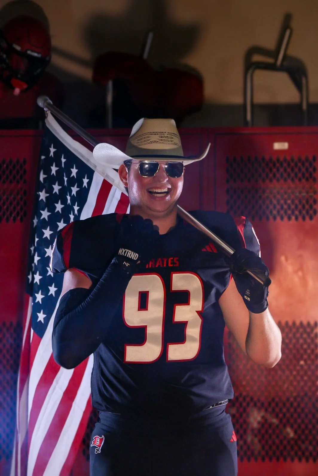 A man wearing a football jersey, sunglasses, a cowboy hat, and gloves is smiling and holding a large American flag while standing in front of red lockers.