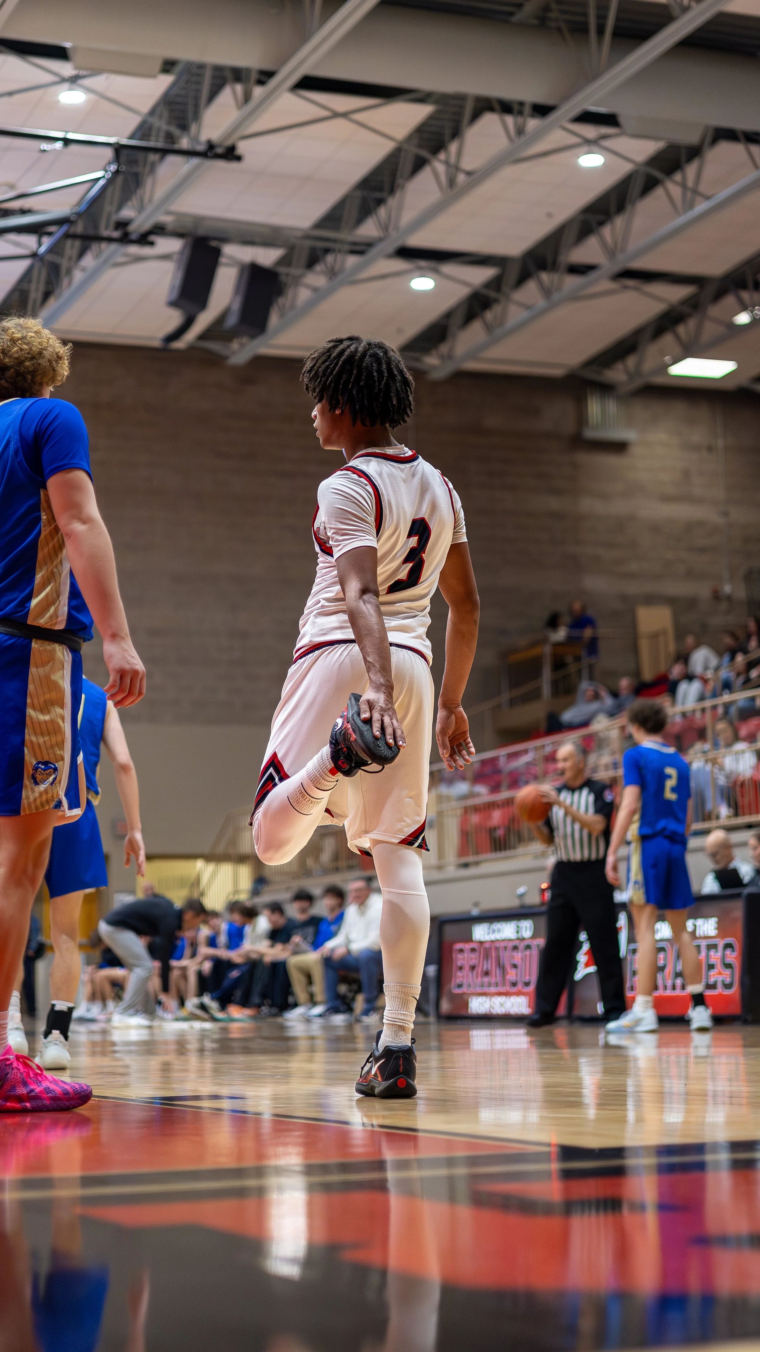 A basketball player in a white jersey stretching on a wooden basketball court in an indoor gym, with other players, a referee, and spectators in the background.
