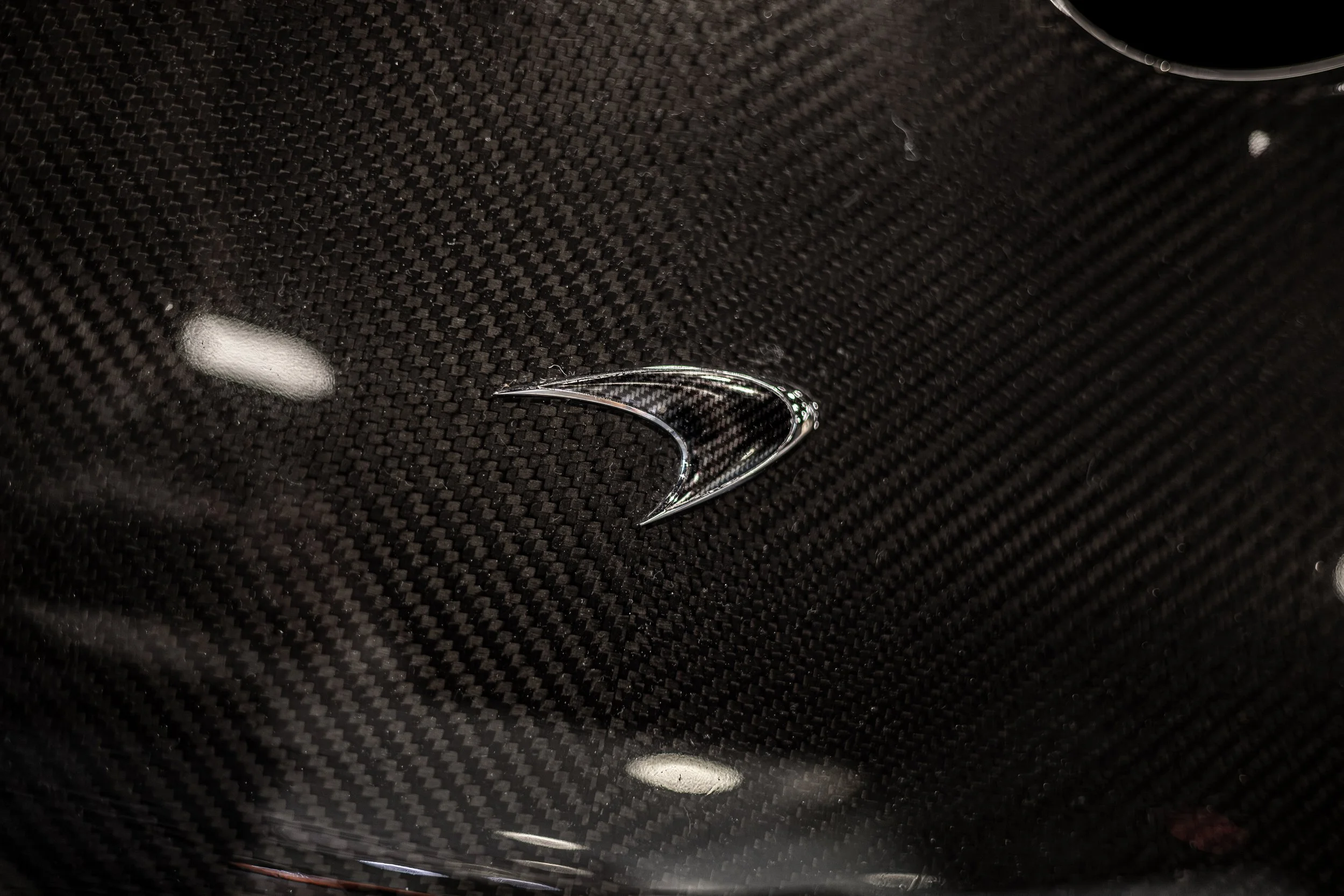 Close-up of a black carbon fiber surface with a chrome logo in the shape of a stylized, curved arrow or swoosh.