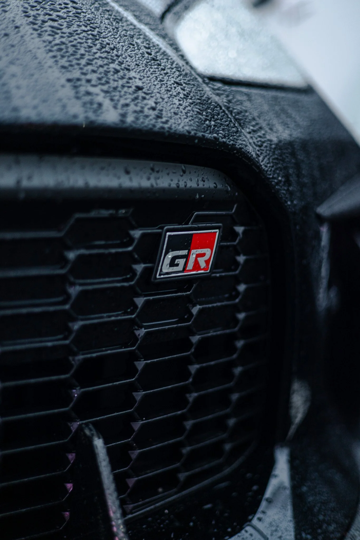 Close-up of a black car grille with a red and silver 'GR' badge, wet from rain.