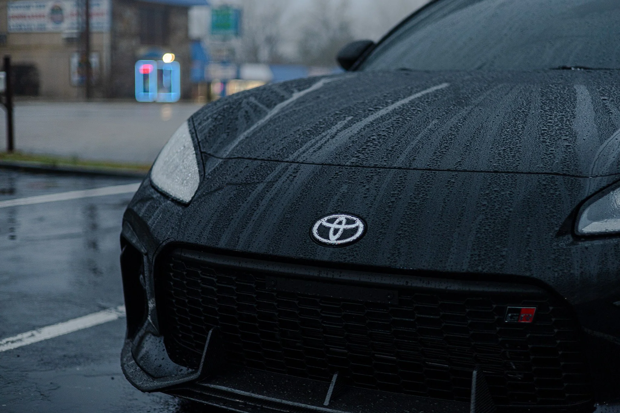 Close-up of a wet black Toyota car in a parking lot, with water droplets on the hood and headlights, during rainy weather.