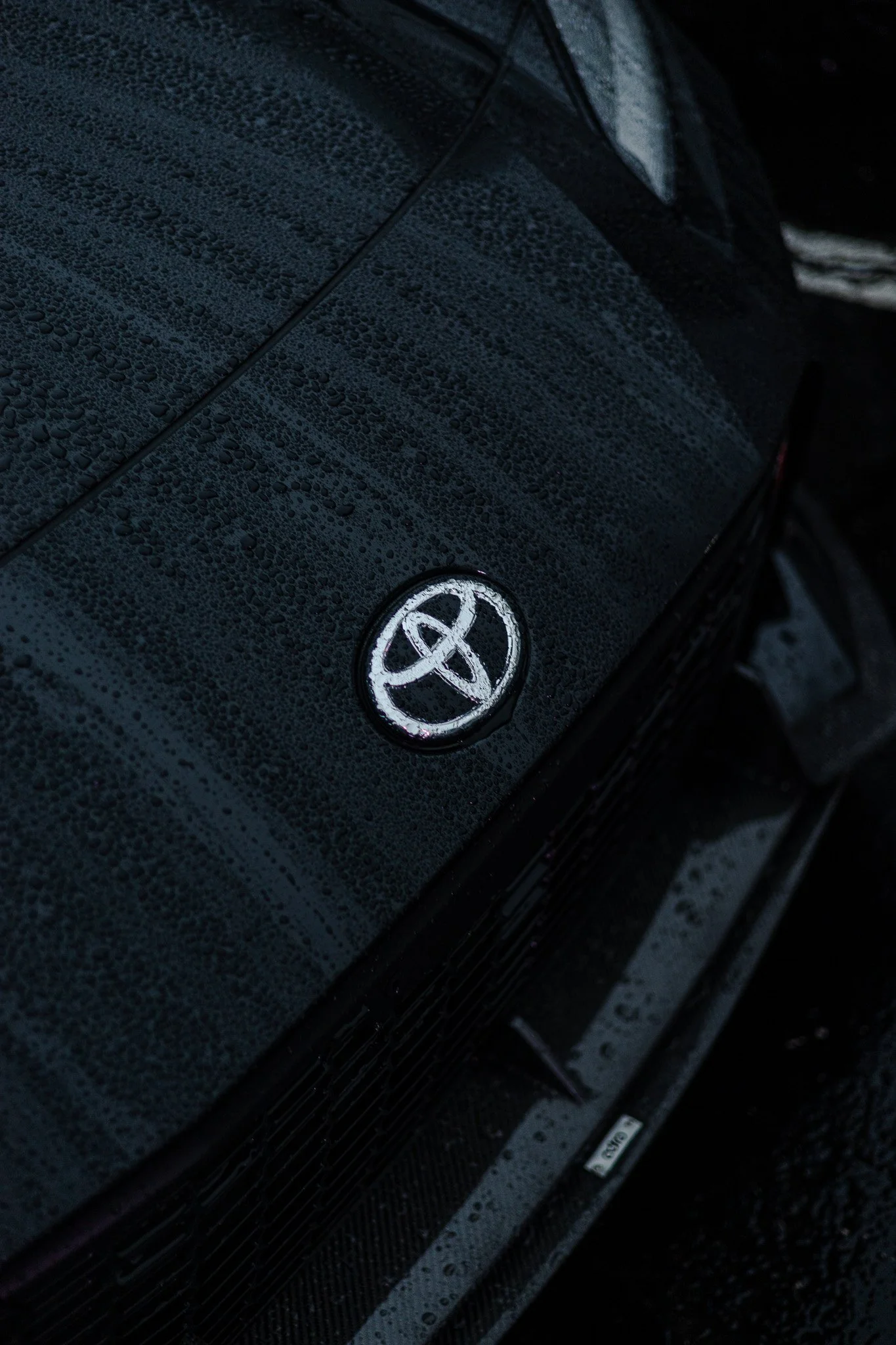 Close-up of a black car's front with water droplets on the hood and an overlapping Toyota logo.