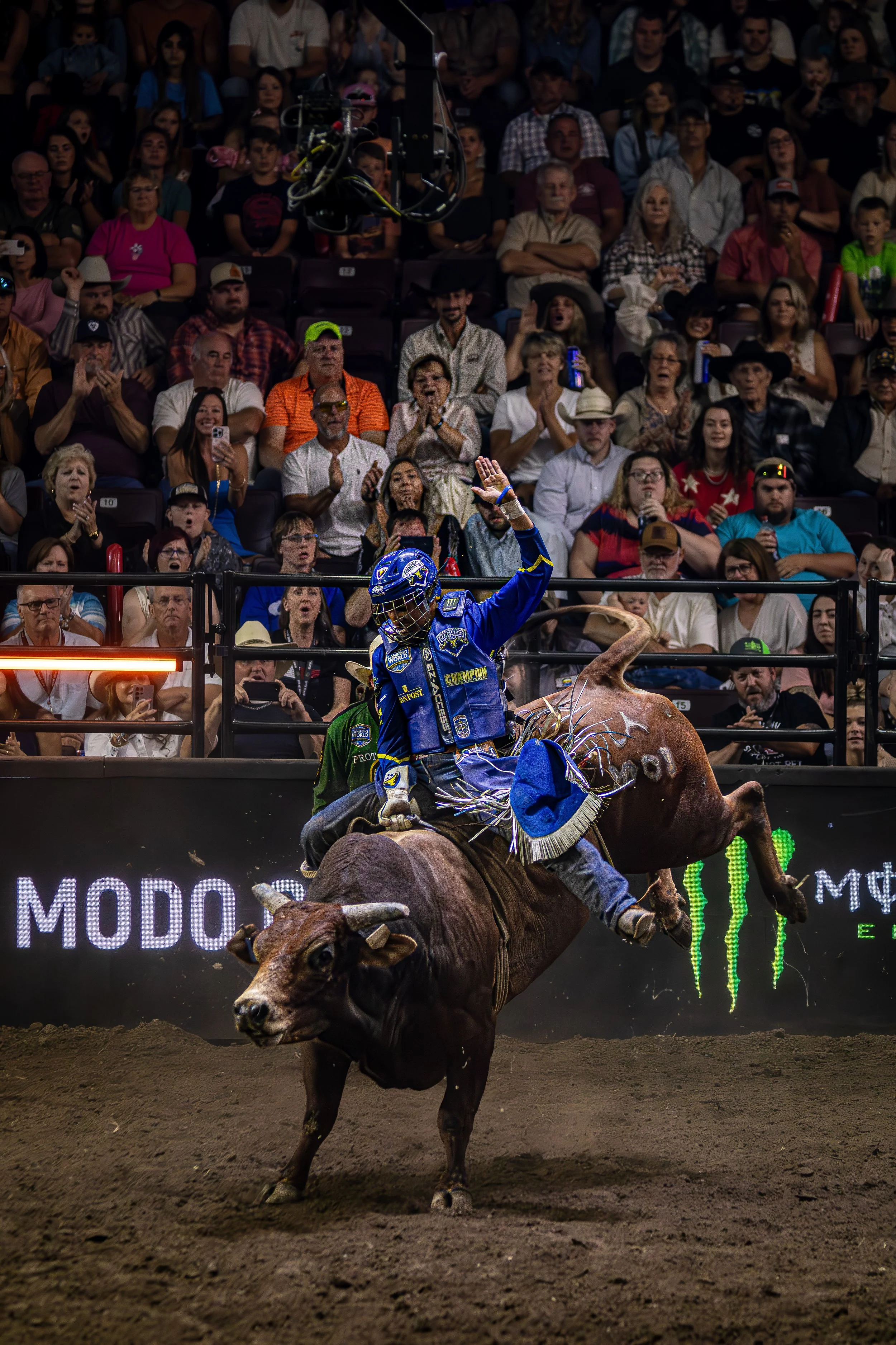 A rodeo cowboy riding a bucking bull in an arena with a large audience watching from the stands.