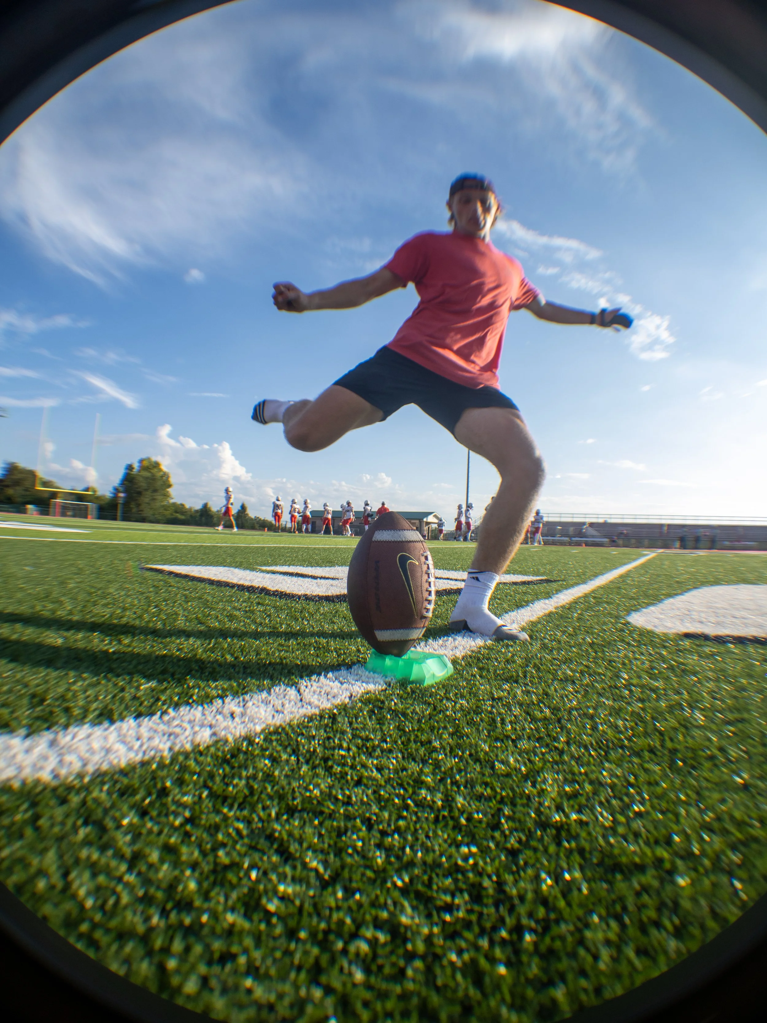 Football player in red shirt and black shorts jumping over football on field during practice.
