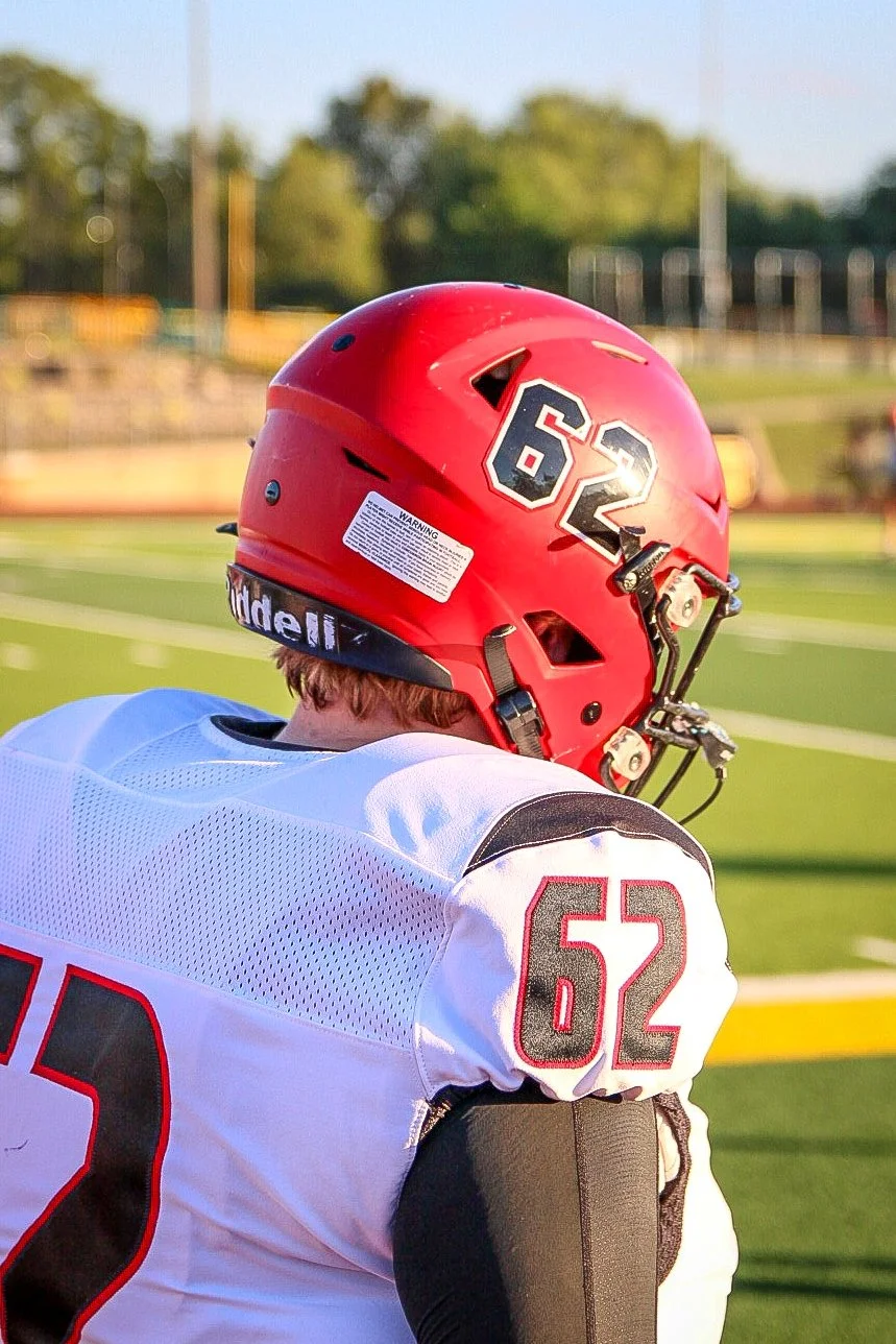 A football player wearing a red helmet with the number 62 on it and a white jersey with the number 62 on the shoulder, standing on a football field during daytime.
