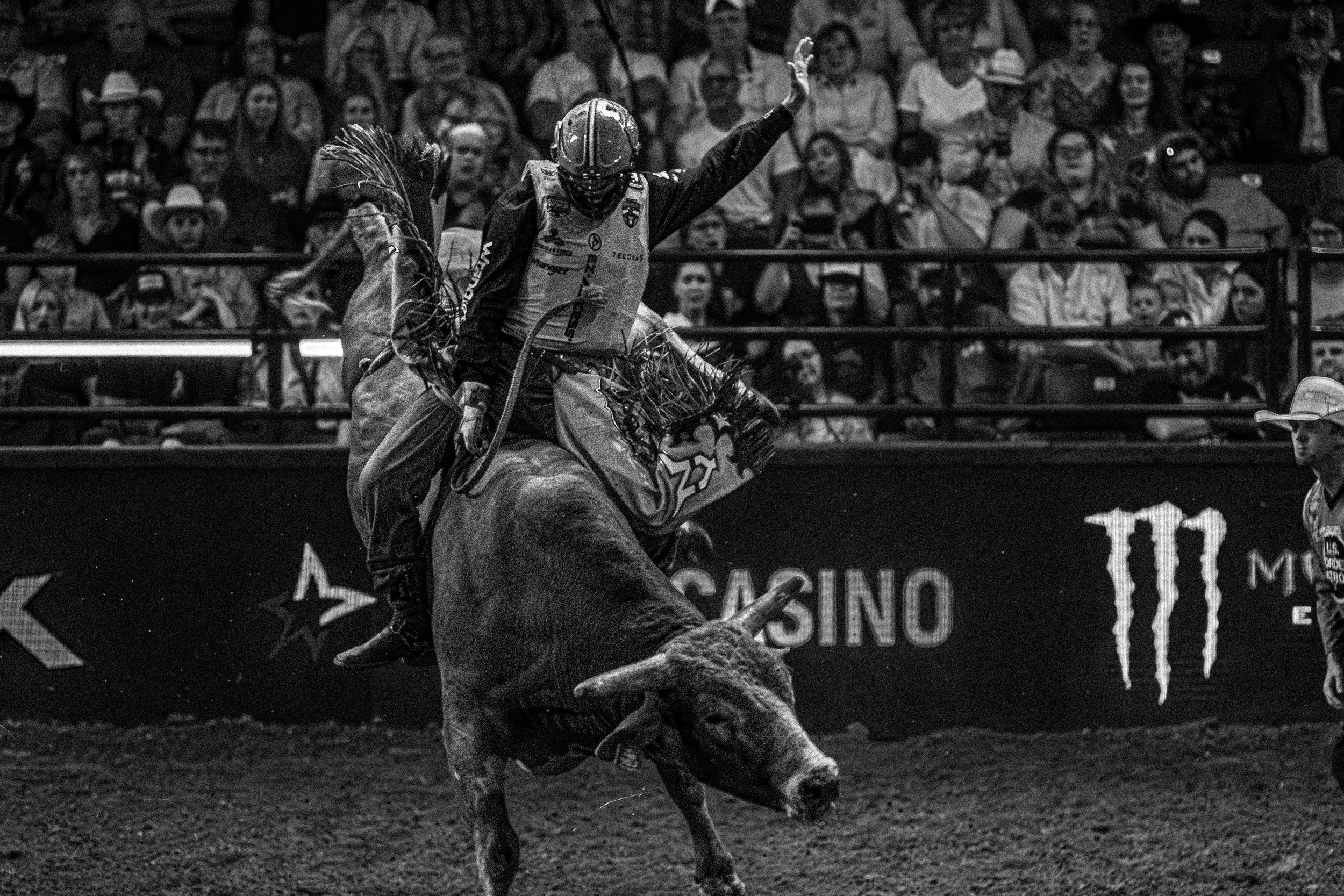 A cowboy riding a bucking bull in a rodeo arena with a crowd watching in the background.