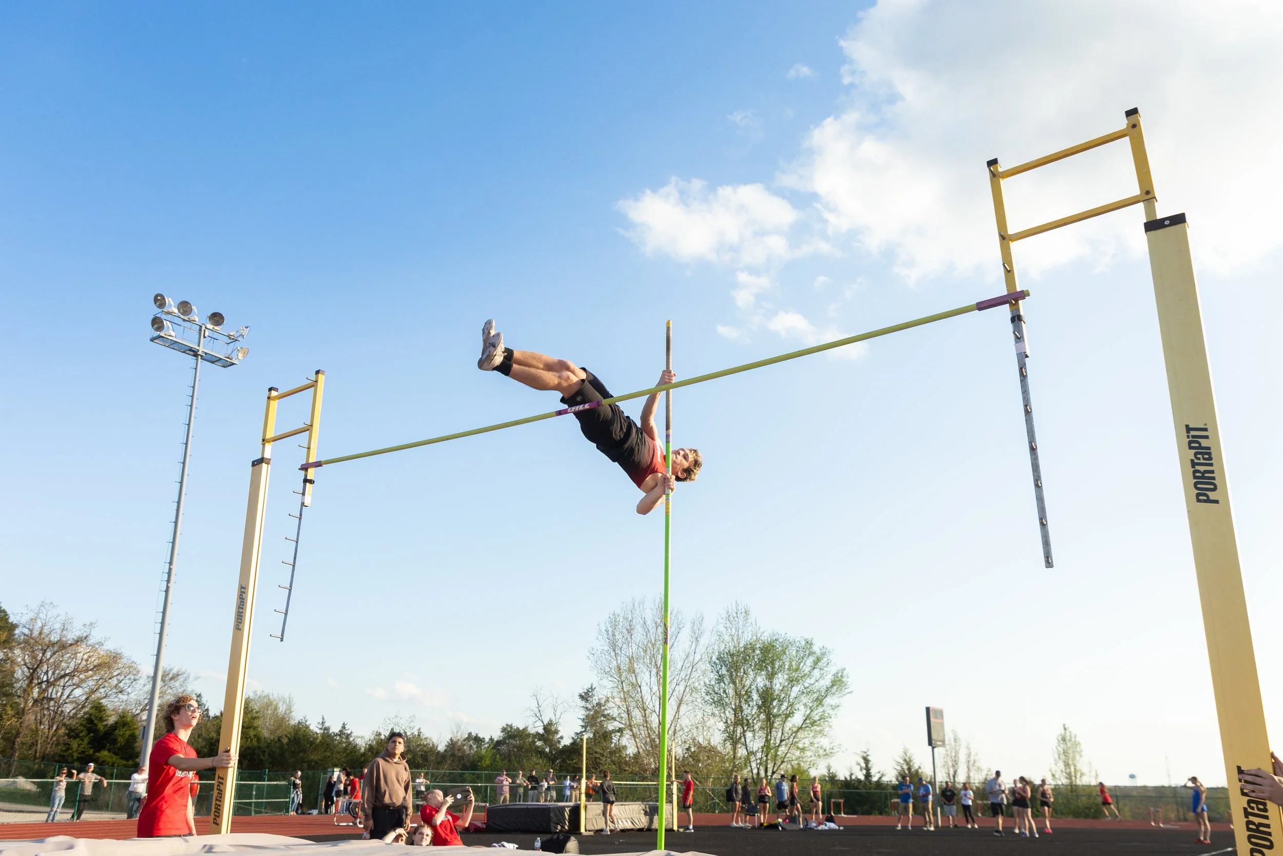 An athlete performing high jump at outdoor track and field event, clearing the bar while holding a pole, with a clear blue sky and spectators in the background.