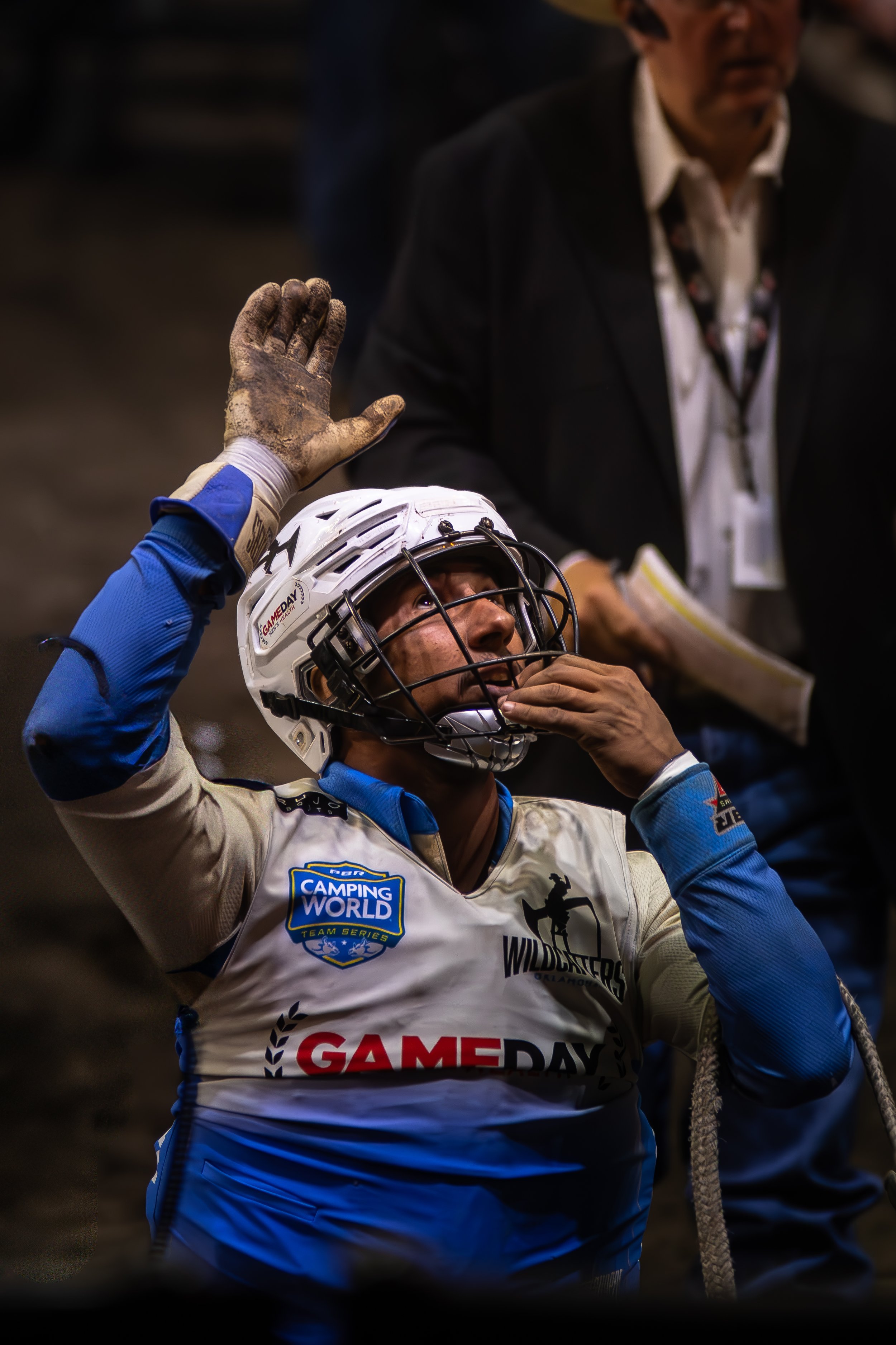 A person in motocross gear with a helmet, raising their right hand and touching their chin in a gesture, while another individual in dark clothing stands nearby holding a script or notes.