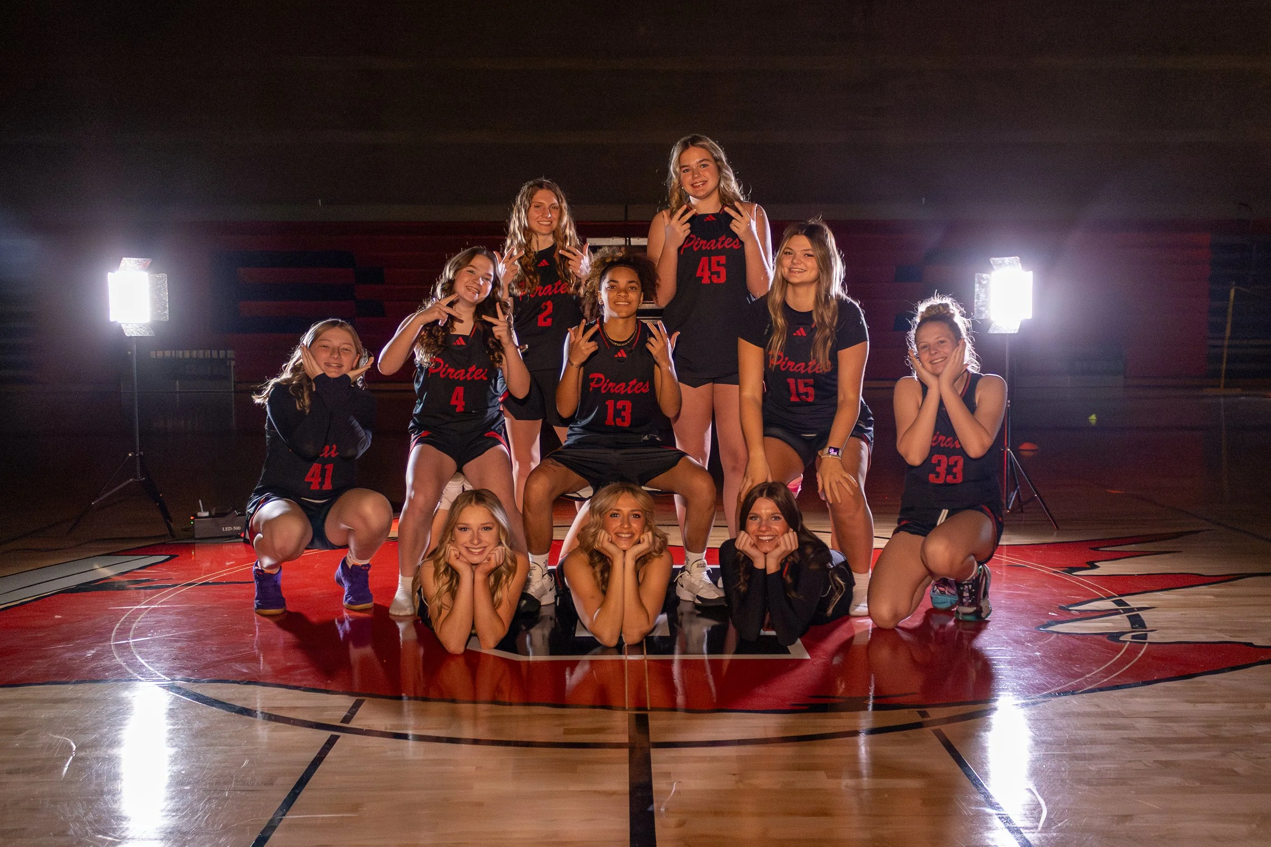 A girls' volleyball team posing on a basketball court with their coach, wearing black jerseys with red lettering and numbers, with bright studio lights behind them.