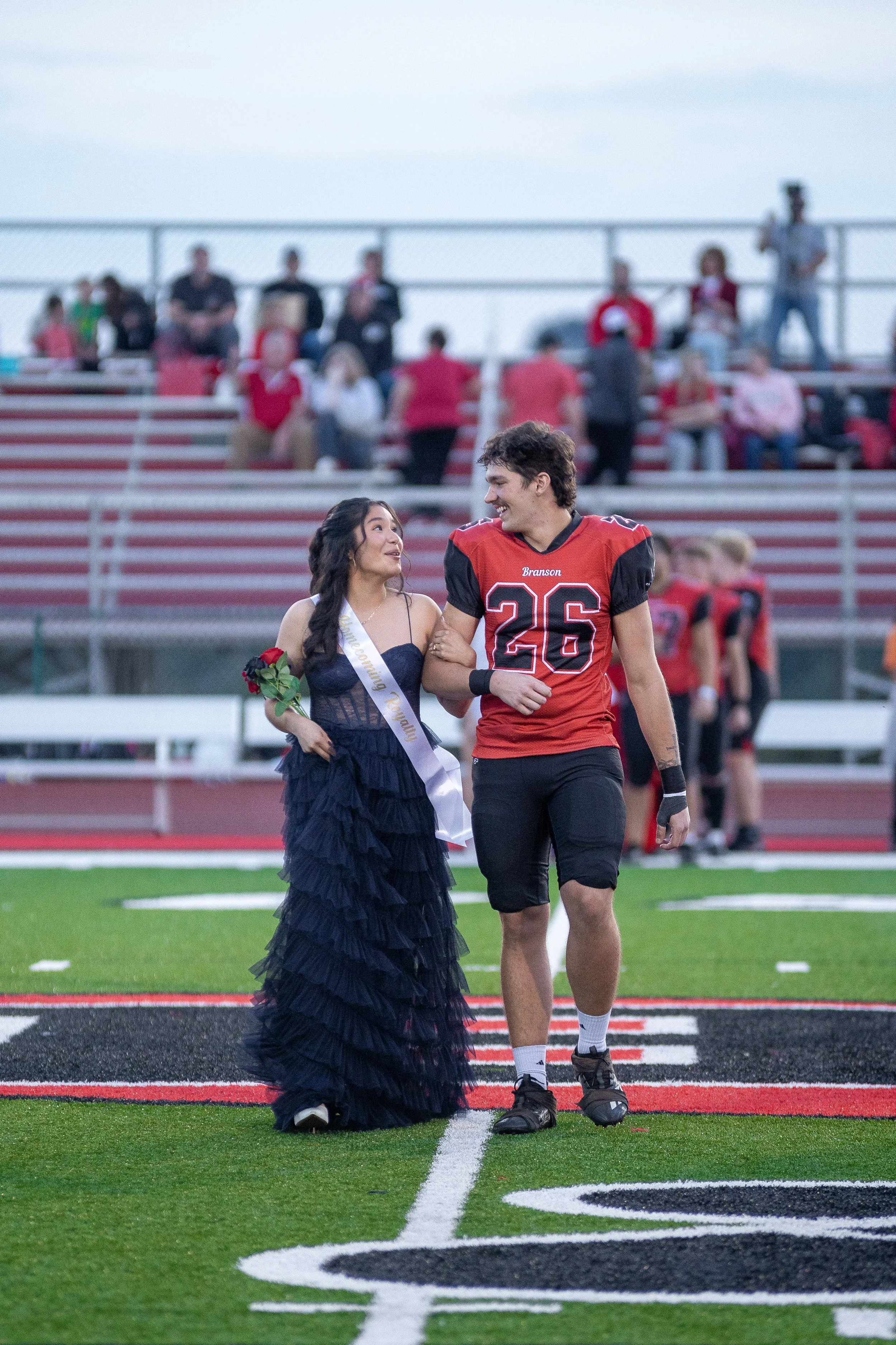 A young woman wearing a sash and holding a red rose walks arm-in-arm with a football player in a red and black uniform on a football field, smiling at each other. Spectators are in the background.