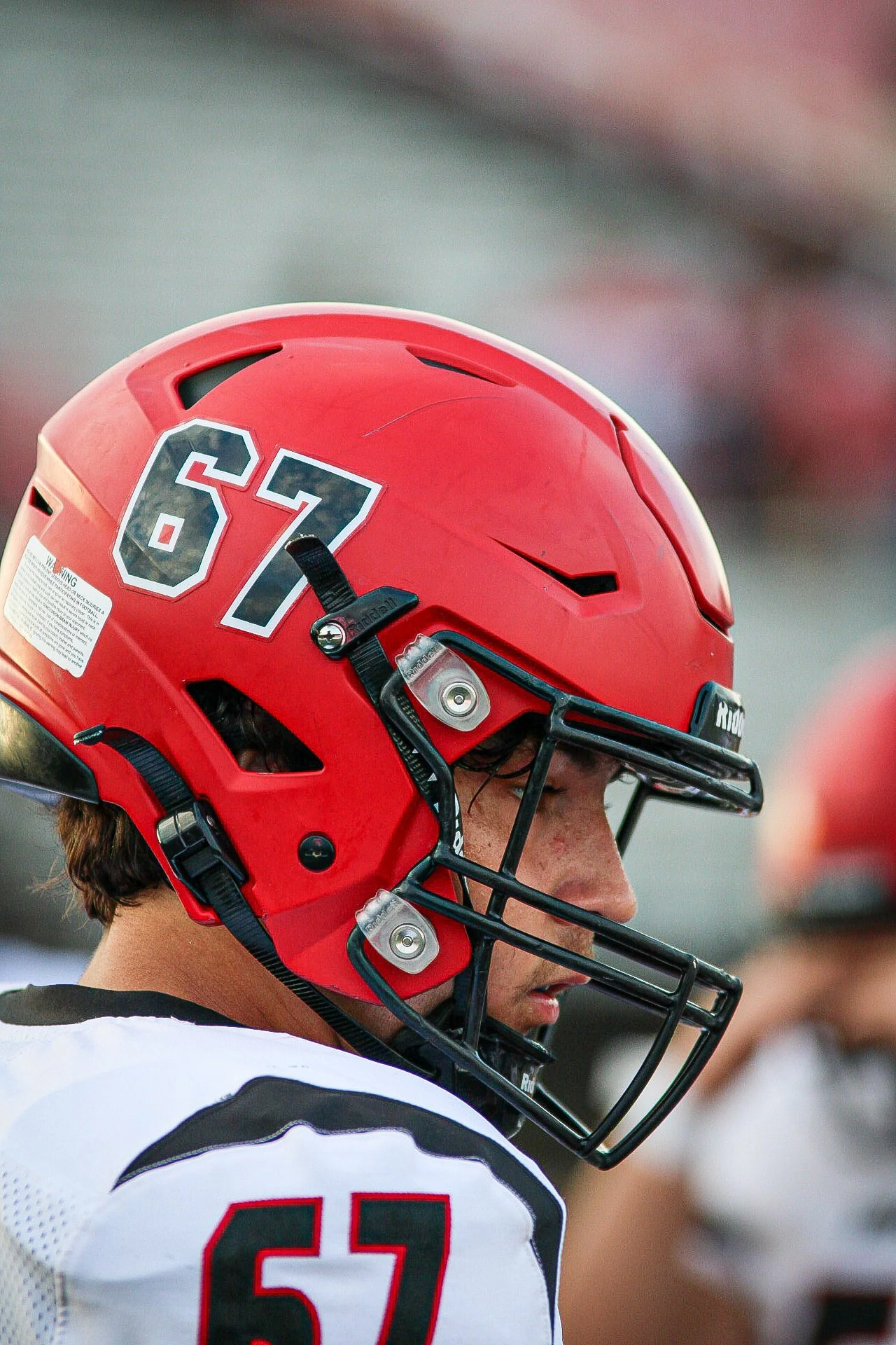 Close-up of a football player wearing a red helmet with the number 67, and a white jersey with black and red accents, on a football field.