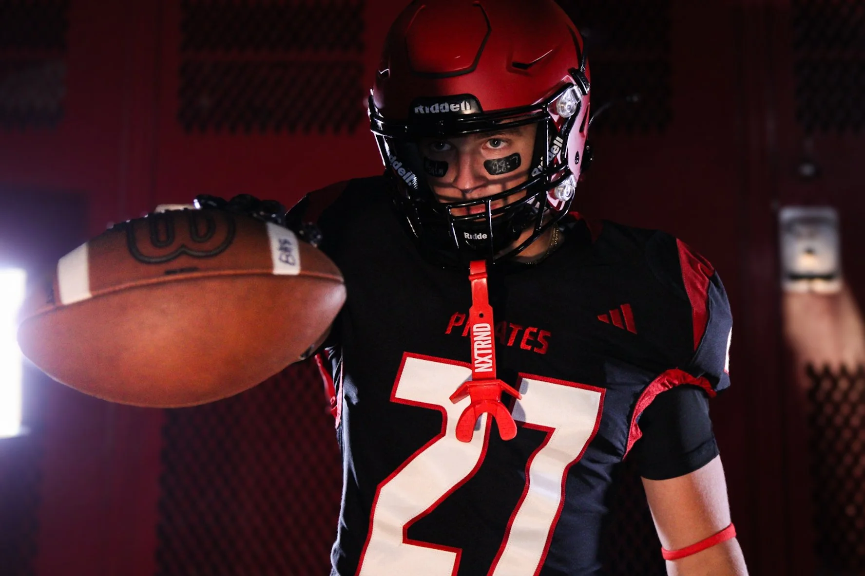 A football player in a helmet and uniform holding a football in a locker room.