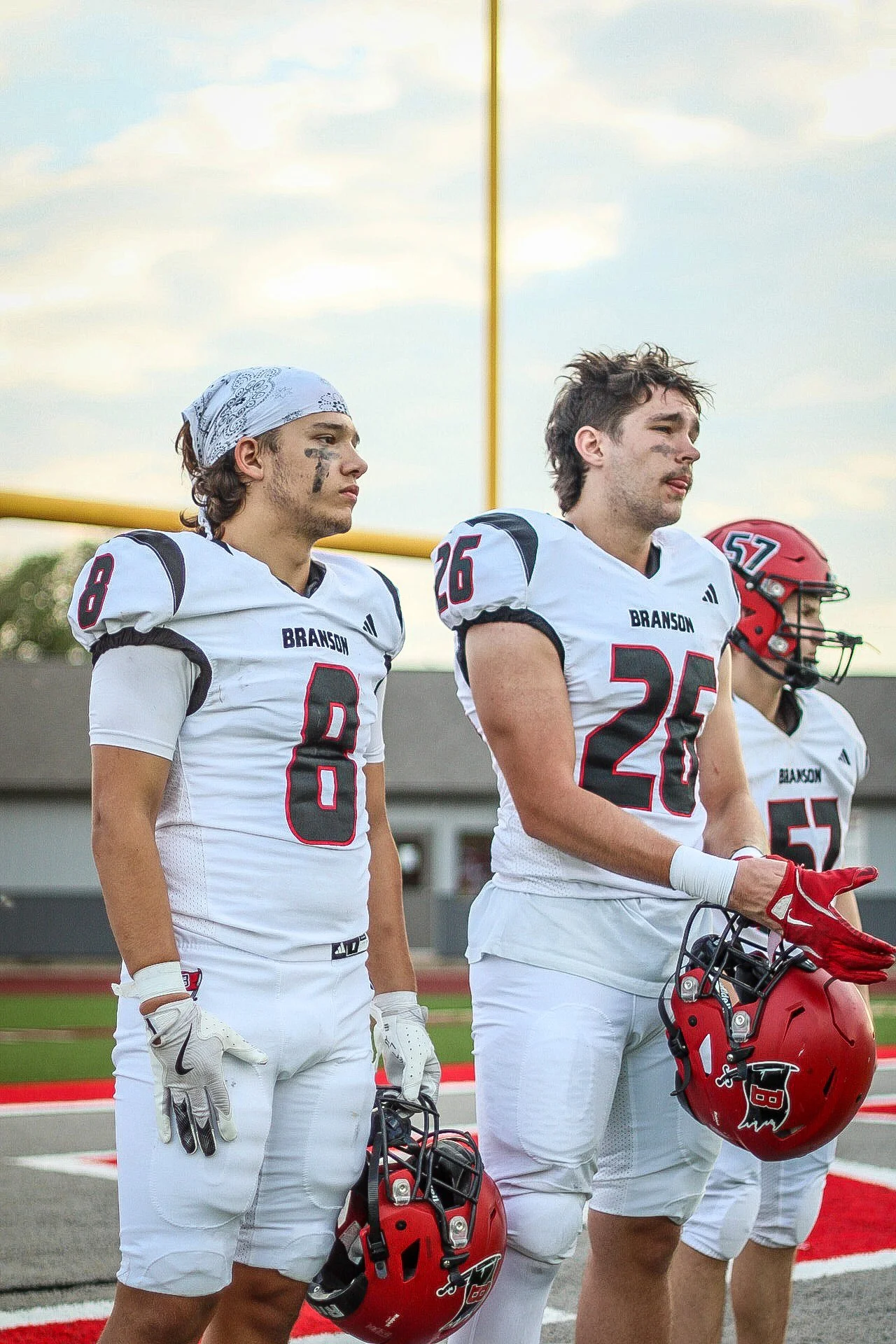Three football players standing on the field during the national anthem, holding their helmets. The players wear white uniforms with black and red accents, and black eye paint. The background shows a football goal post and a partly cloudy sky.