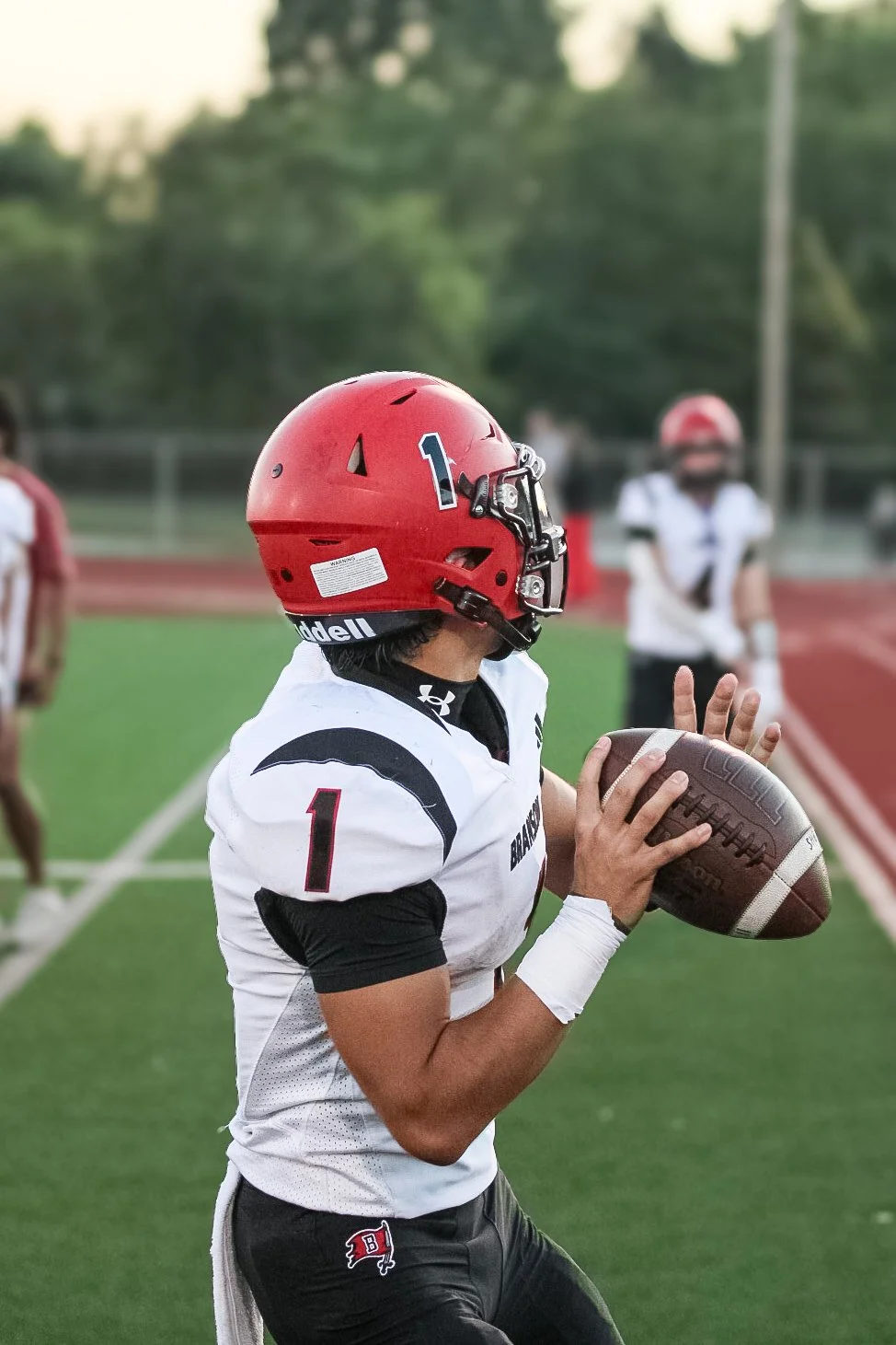 Football player in a white uniform and red helmet holding a football on a field with other players in the background.