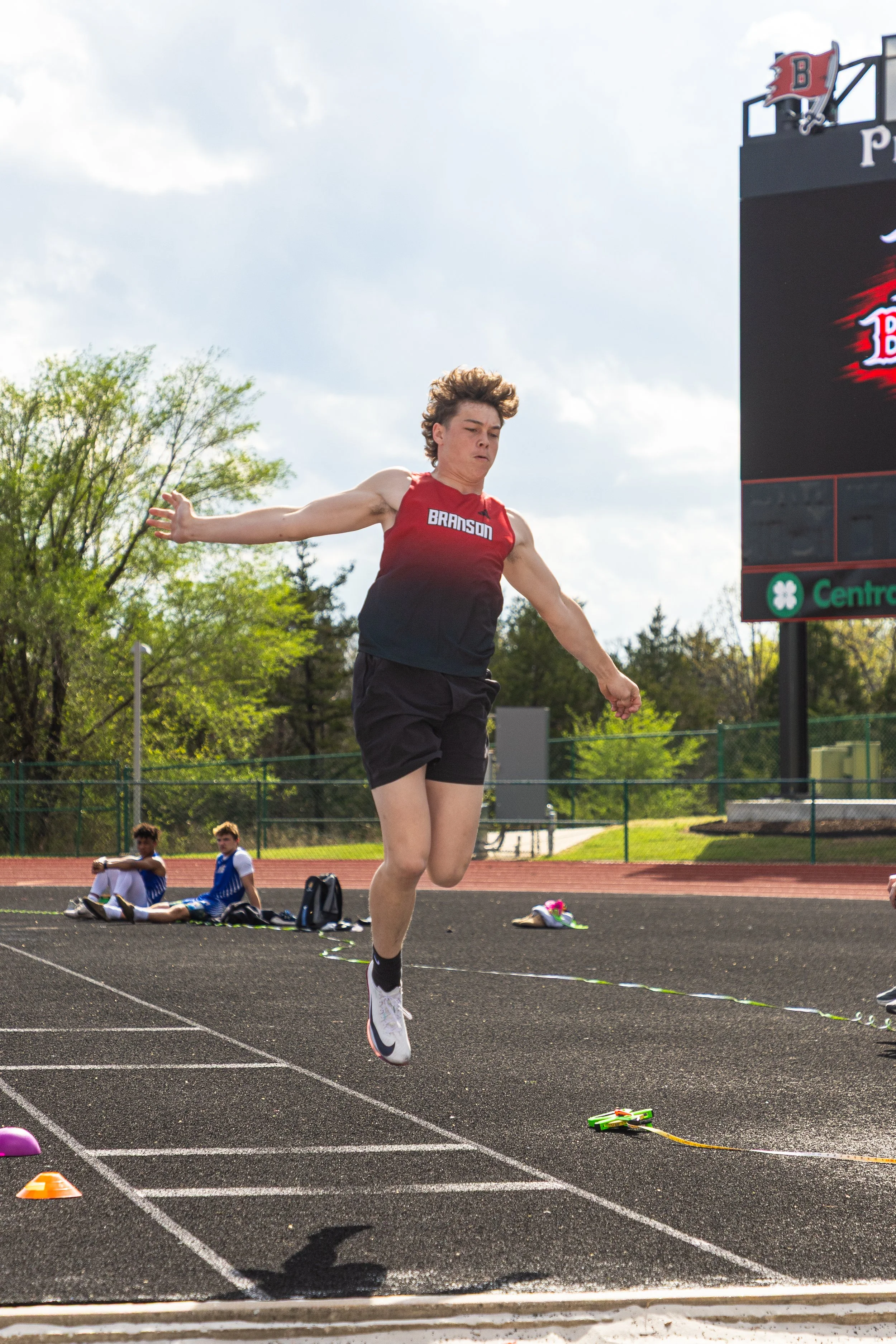 A male athlete in a red and black uniform jumps over a hurdle on a track during a track and field event, with a scoreboard and other athletes sitting on the sidelines in the background.