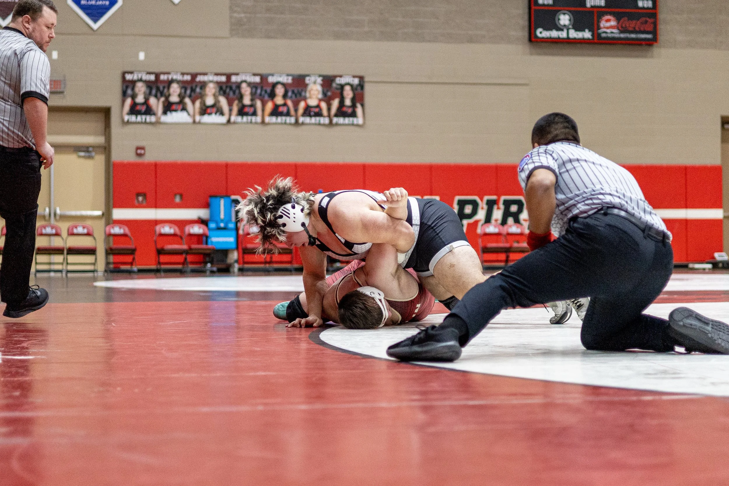 A female wrestler in a black and pink singlet and white protective headgear is pinning a male wrestler in a maroon singlet on a red wrestling mat, with a referee kneeling nearby and observing closely.