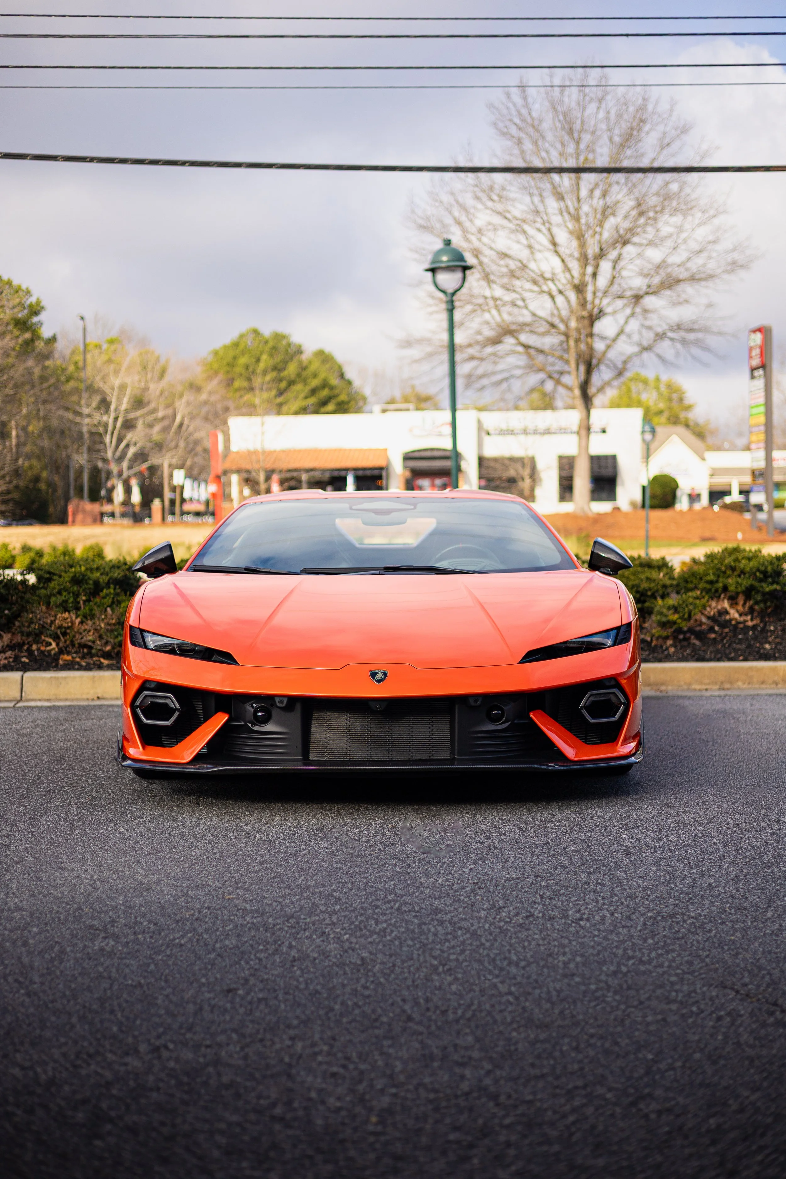 Red Lamborghini sports car parked on asphalt in front of a shopping center with trees and cloudy sky in the background.