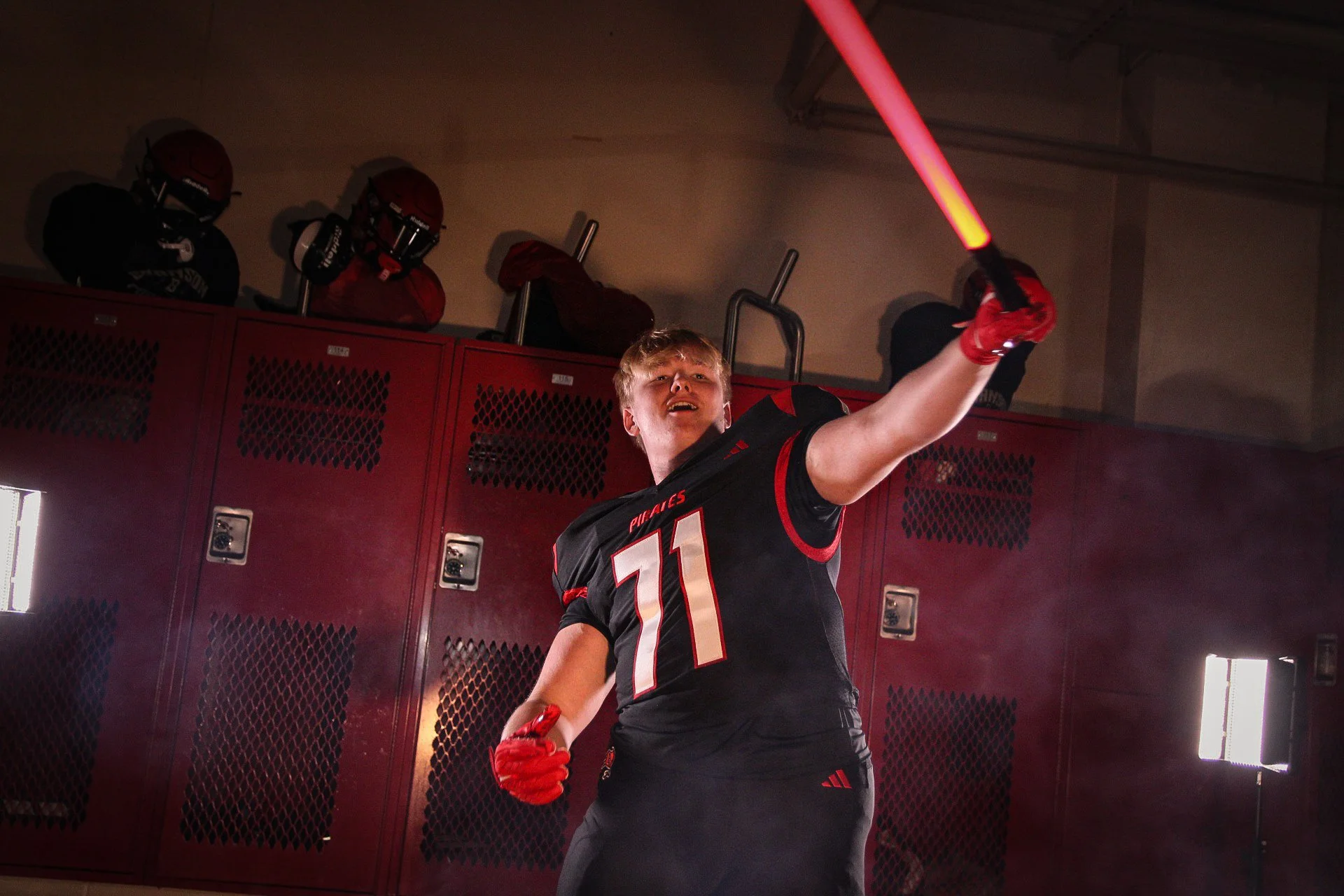 A young football player in a black and red uniform holding a red light saber in a locker room.