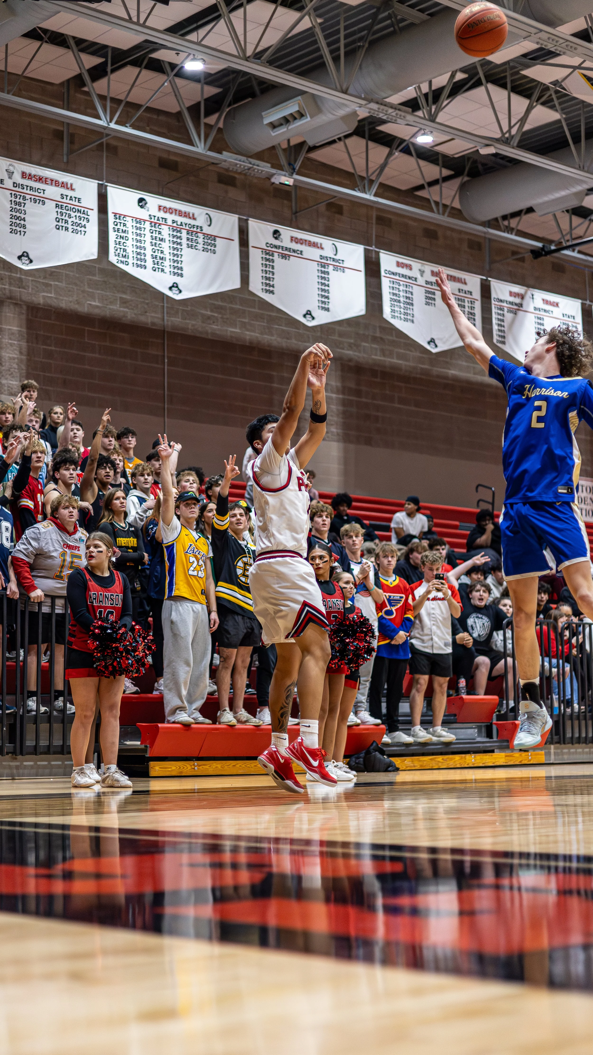 A basketball game with a player in a blue jersey jumping to shoot while another player in a white jersey attempts to block. A large crowd of spectators and cheerleaders are watching, with many standing and raising their hands. The game is taking plac