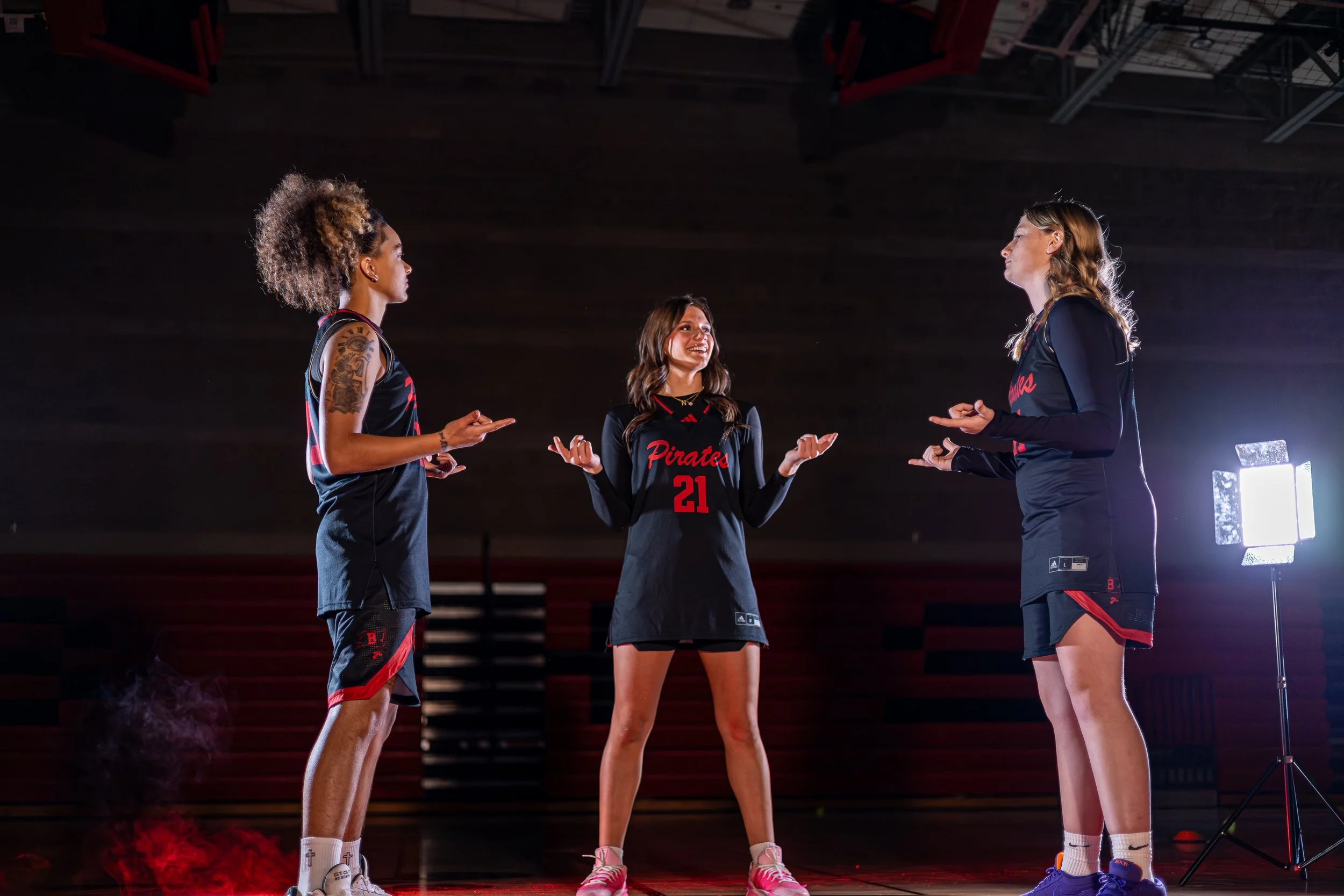 Three female athletes in black volleyball uniforms standing on a basketball court, engaging in conversation. One is standing in the middle with the number 21, and the other two are facing her, with one on each side. Bright lighting and a dark backgro