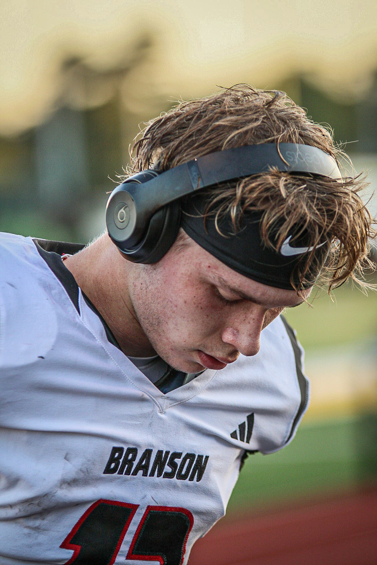 A young male football player wearing a white jersey with 'BRANSON' and number 10, black headband, and black Beats headphones, looking down during a game or practice.