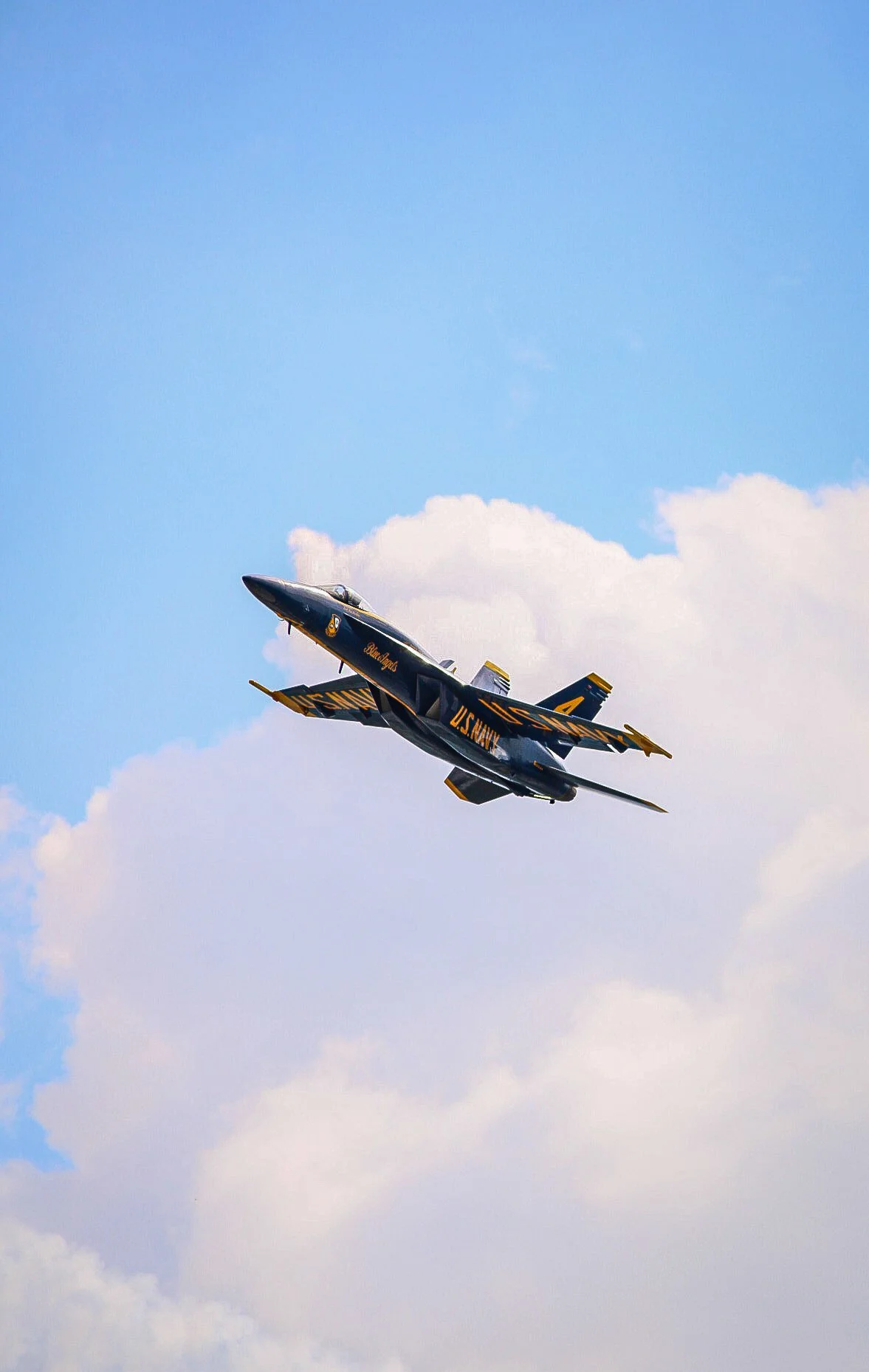 A fighter jet with U.S. Navy markings flying in the sky with scattered clouds.
