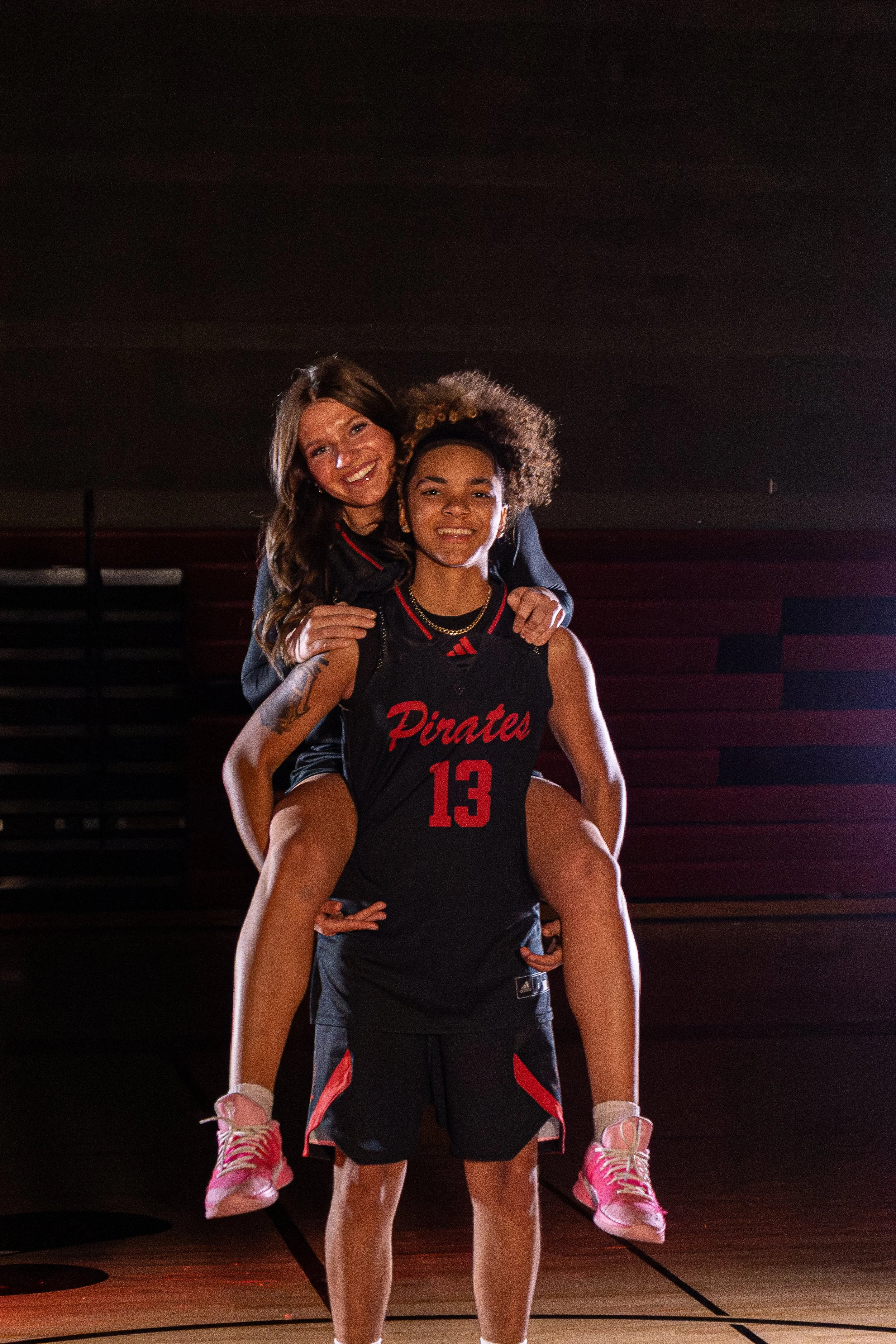 A woman giving a piggyback ride to a young girl in a black basketball uniform with red letters and the number 13, on a basketball court at night.