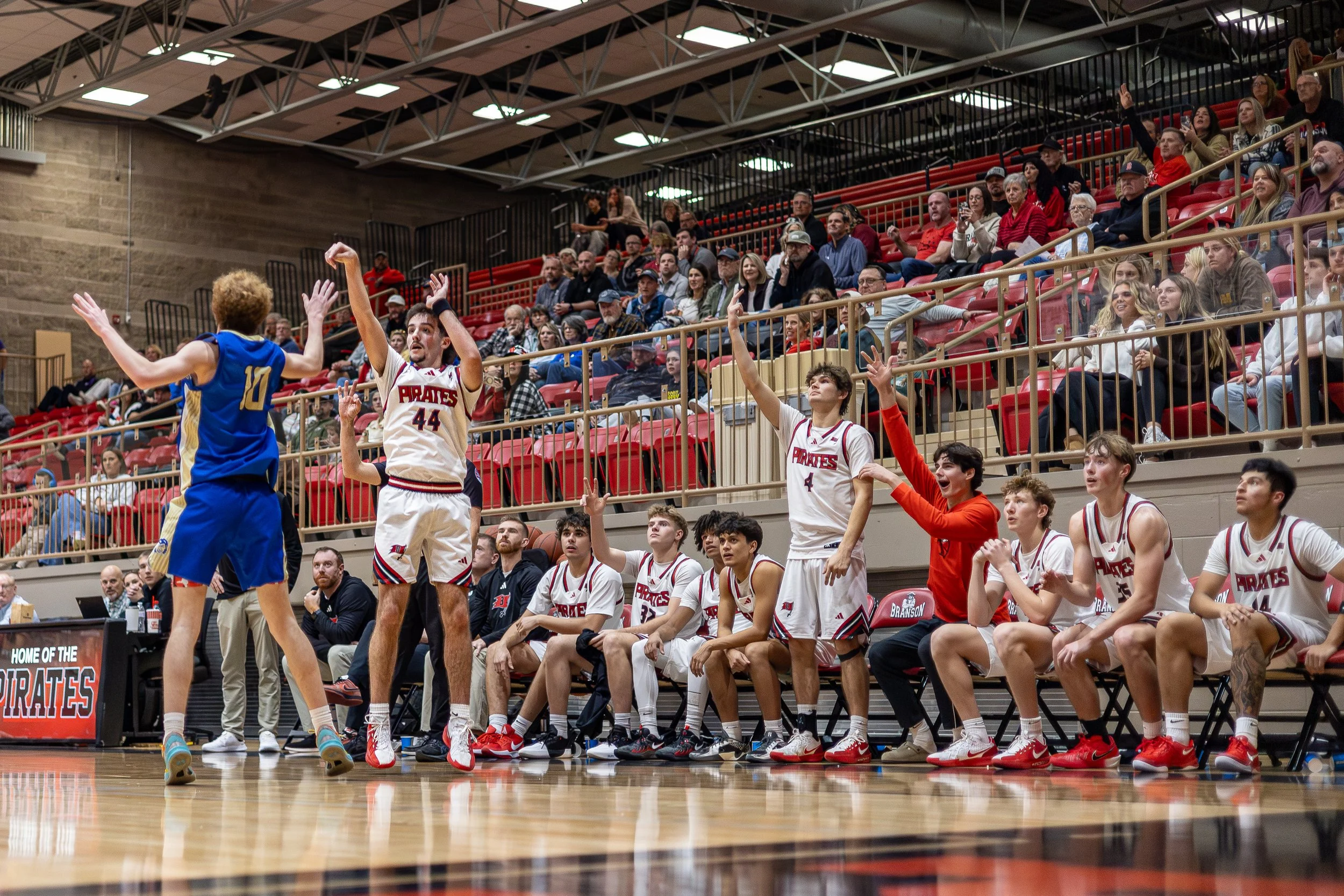 A basketball game with players on the court and spectators in the stands. A player in a blue jersey is leaping with arms extended, facing players in white jerseys from the PRAIES team, who are reacting with raised hands and some seated on the bench. 